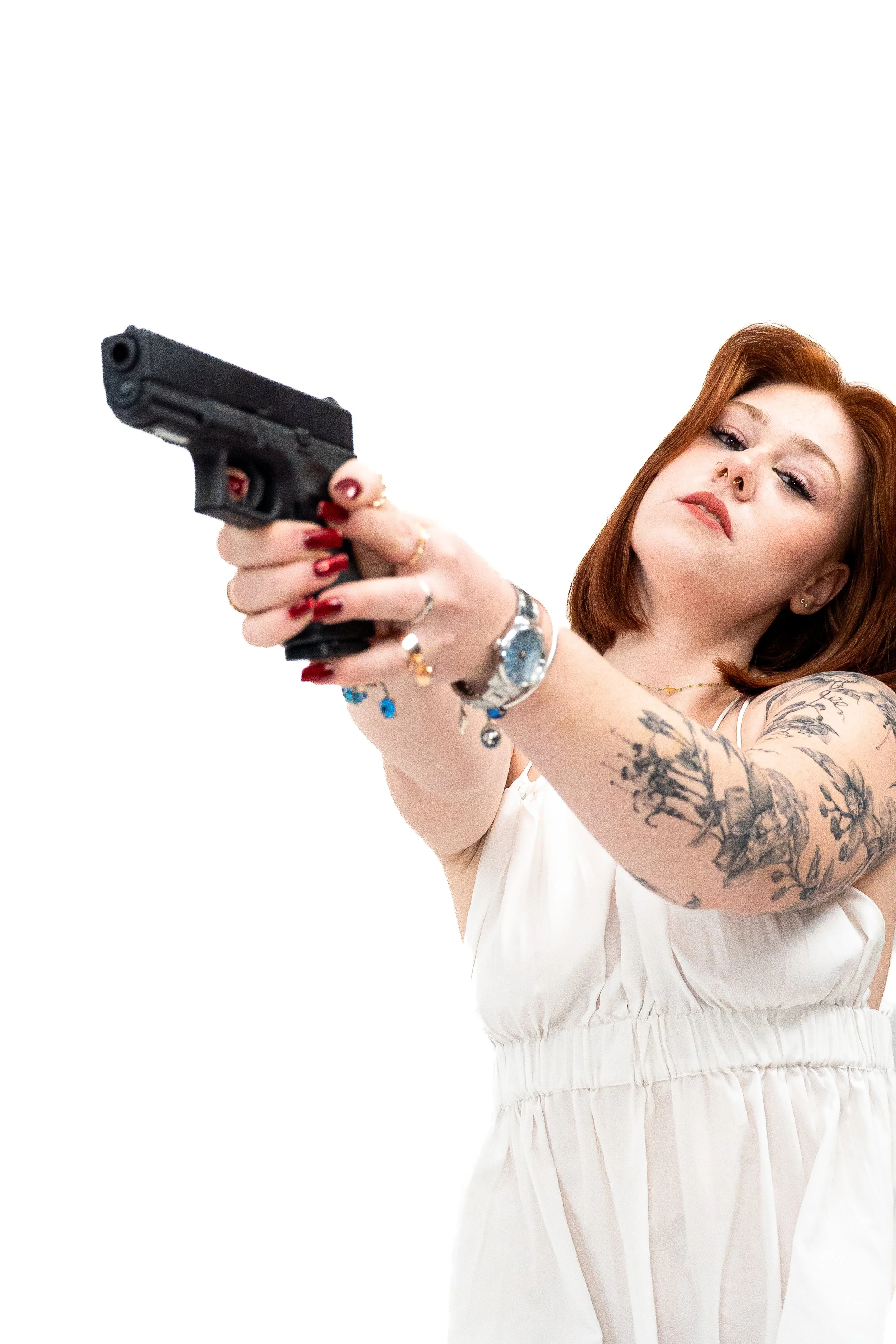 Une femme aux cheveux roux, arborant des tatouages et portant une robe blanche, pointe un pistolet vers l'objectif.