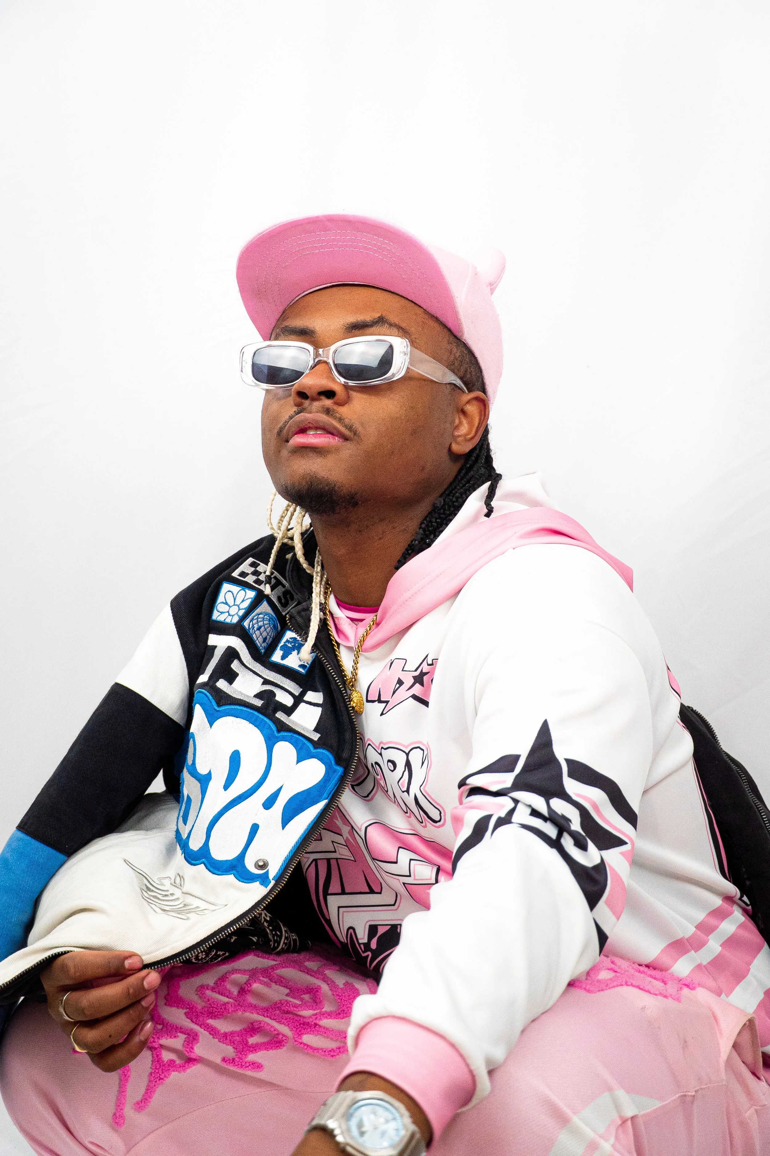 Jeune homme portant une veste colorée, un chapeau rose, des lunettes de soleil, et une chaîne en or, posant contre un fond blanc.