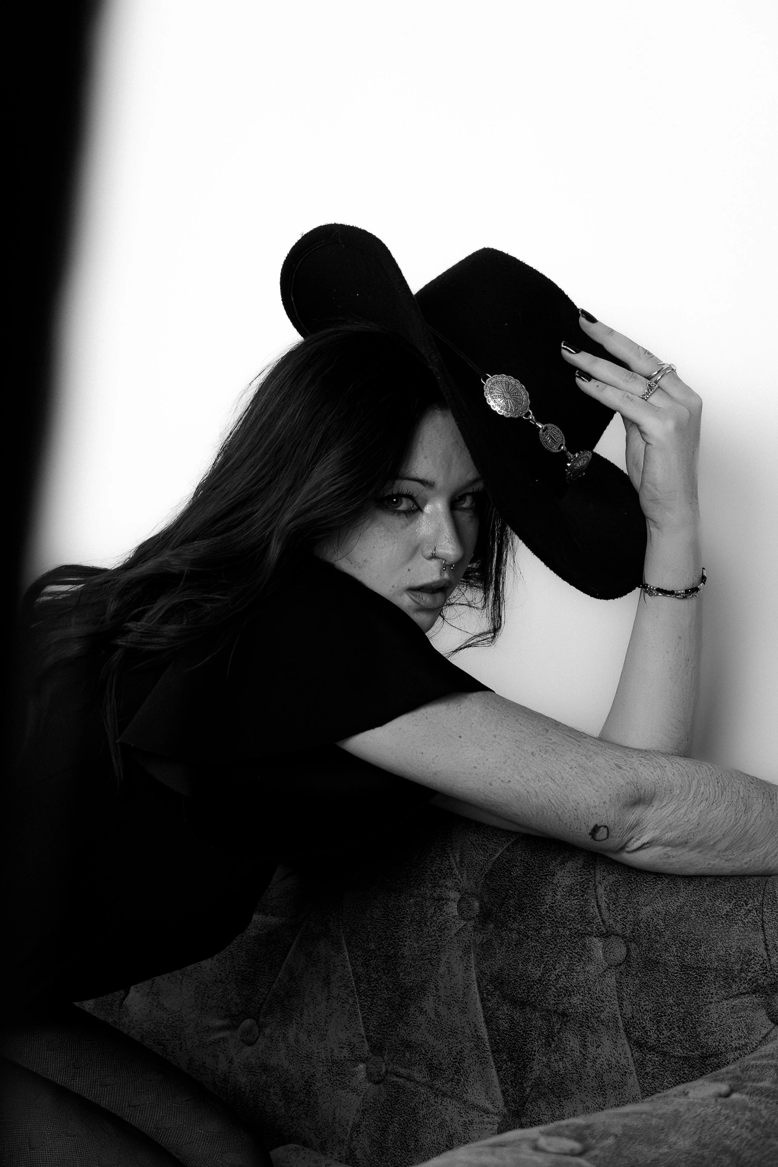Une femme avec des cheveux longs et foncés, portant un chapeau noir, pose sur un canapé en cuir, regardant intensément vers l'objectif.