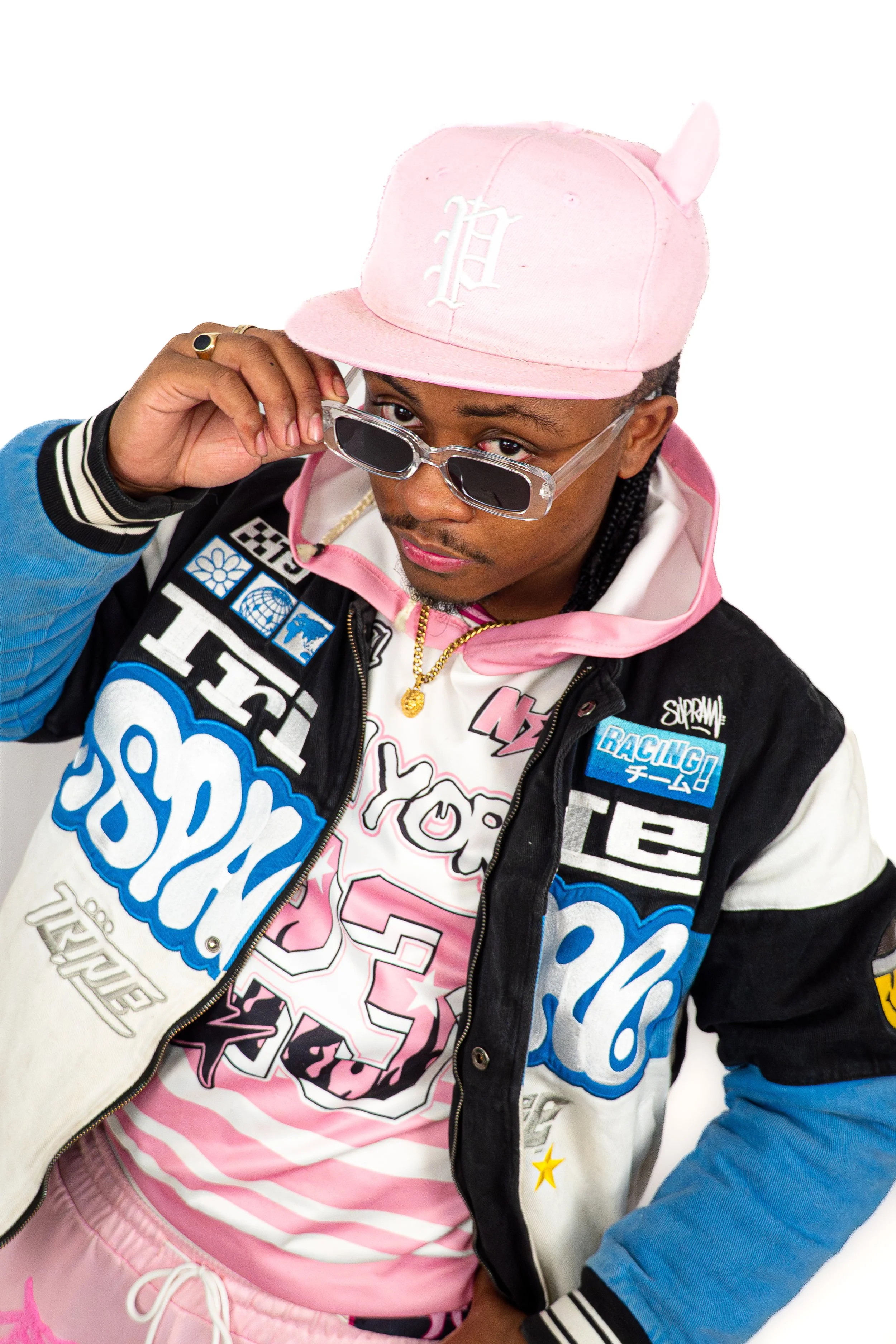 Un homme portant un sweat à capuche coloré, une veste à patchs, un chapeau rose et des lunettes de soleil élégantes, posant avec confiance.