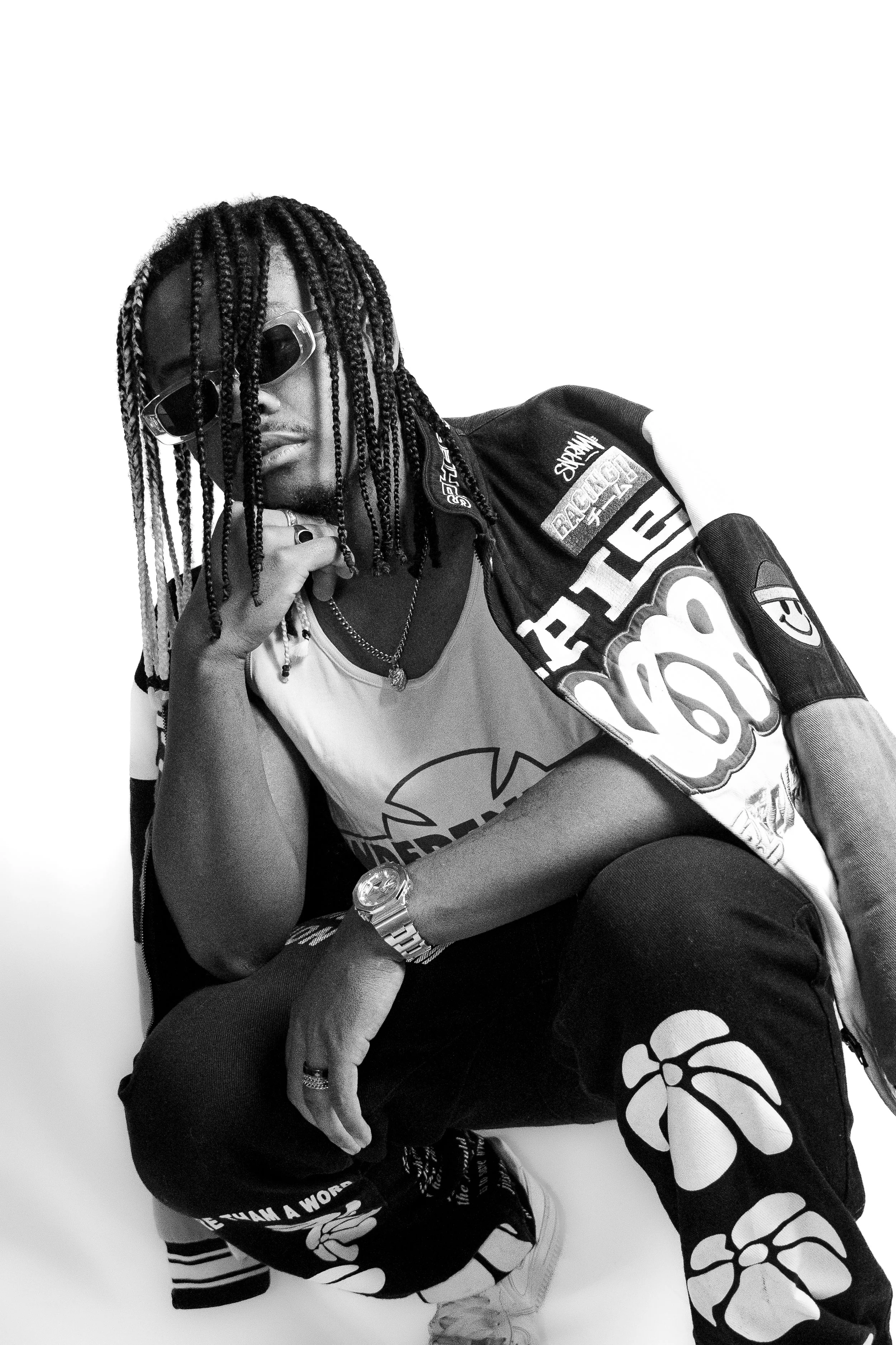 Un homme posant avec des vêtements streetwear, portant des lunettes de soleil, des dreadlocks et des accessoires comme une montre et des bijoux, sur un fond blanc.