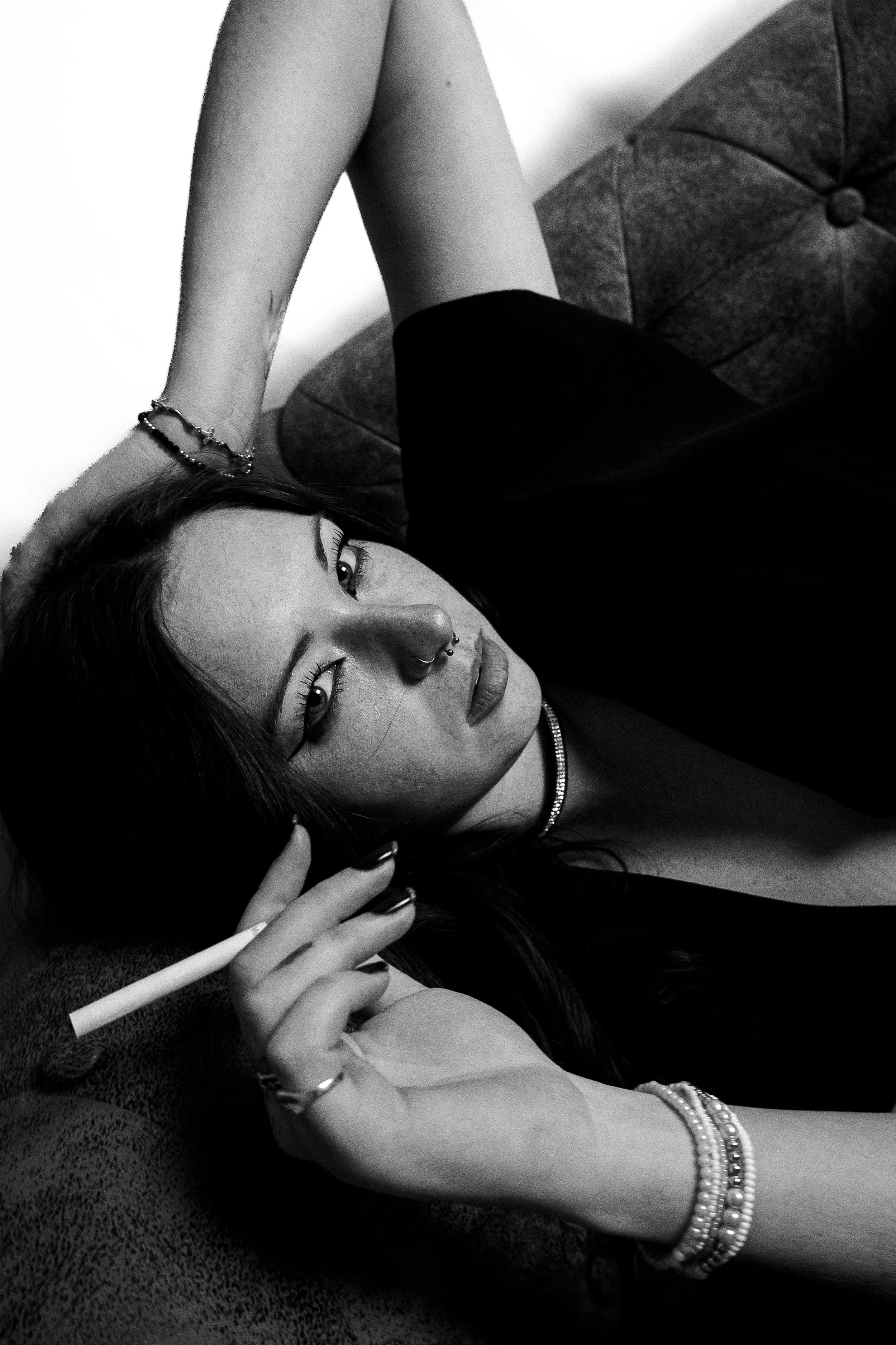 Femme allongée sur un canapé, tenant une cigarette, portant des bijoux, portrait en noir et blanc.