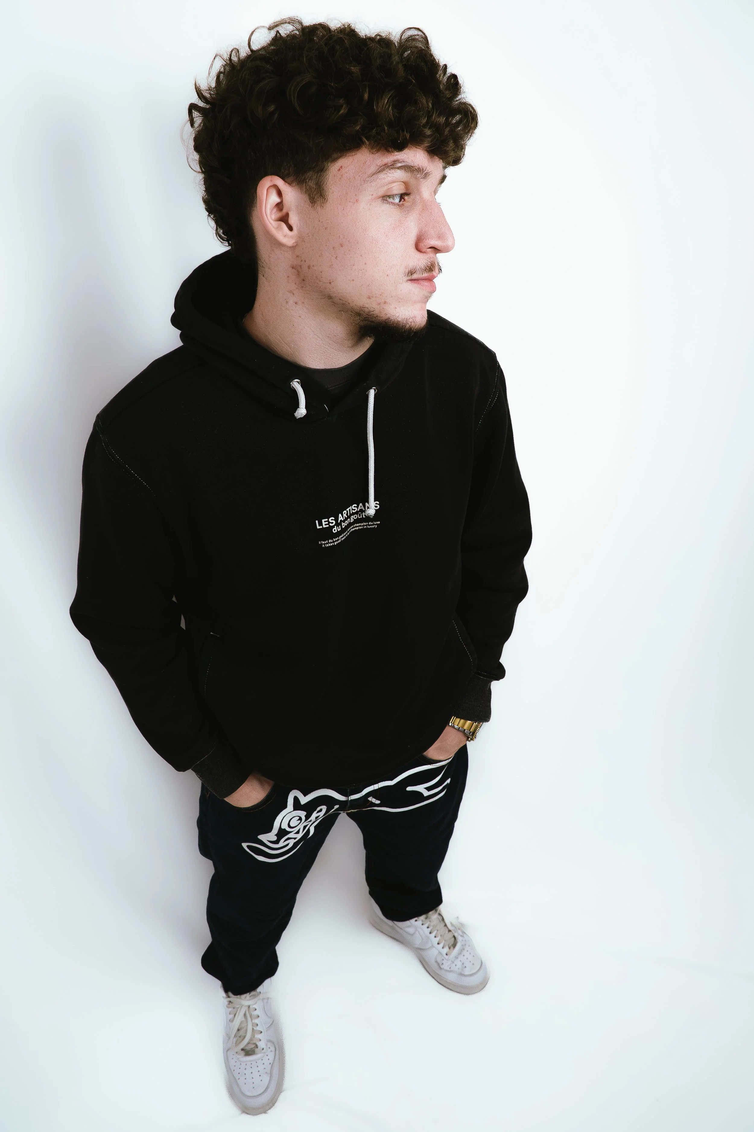 Un jeune homme avec des cheveux bouclés et une barbe porte un sweat noir avec capuche, des pantalons noirs avec un dessin blanc, des baskets blanches et une montre en or. Il se tient contre un mur blanc, regardant vers la droite.