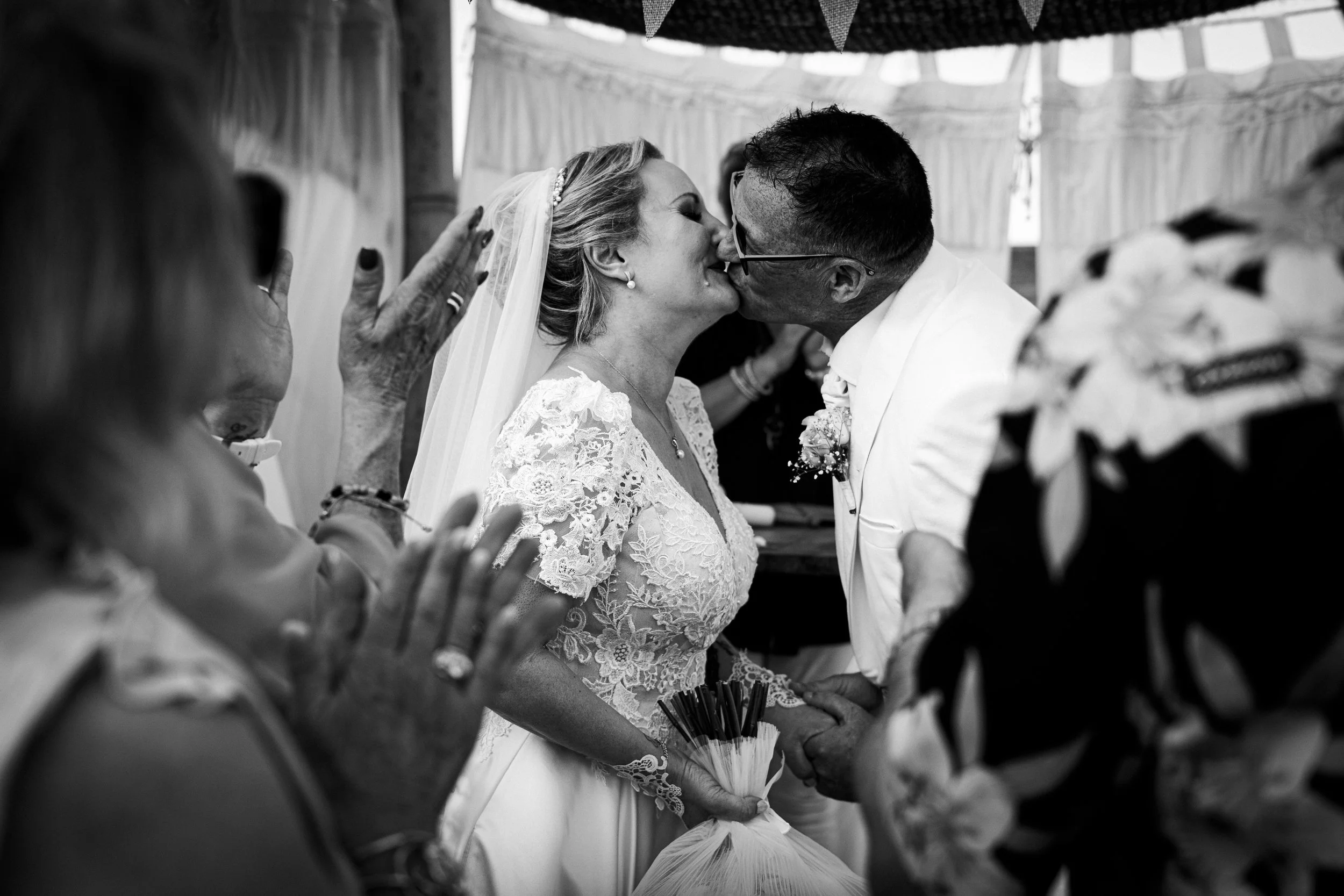 Un couple de mariés s'embrassent lors de leur mariage, entourés de proches, dans une ambiance joyeuse.
