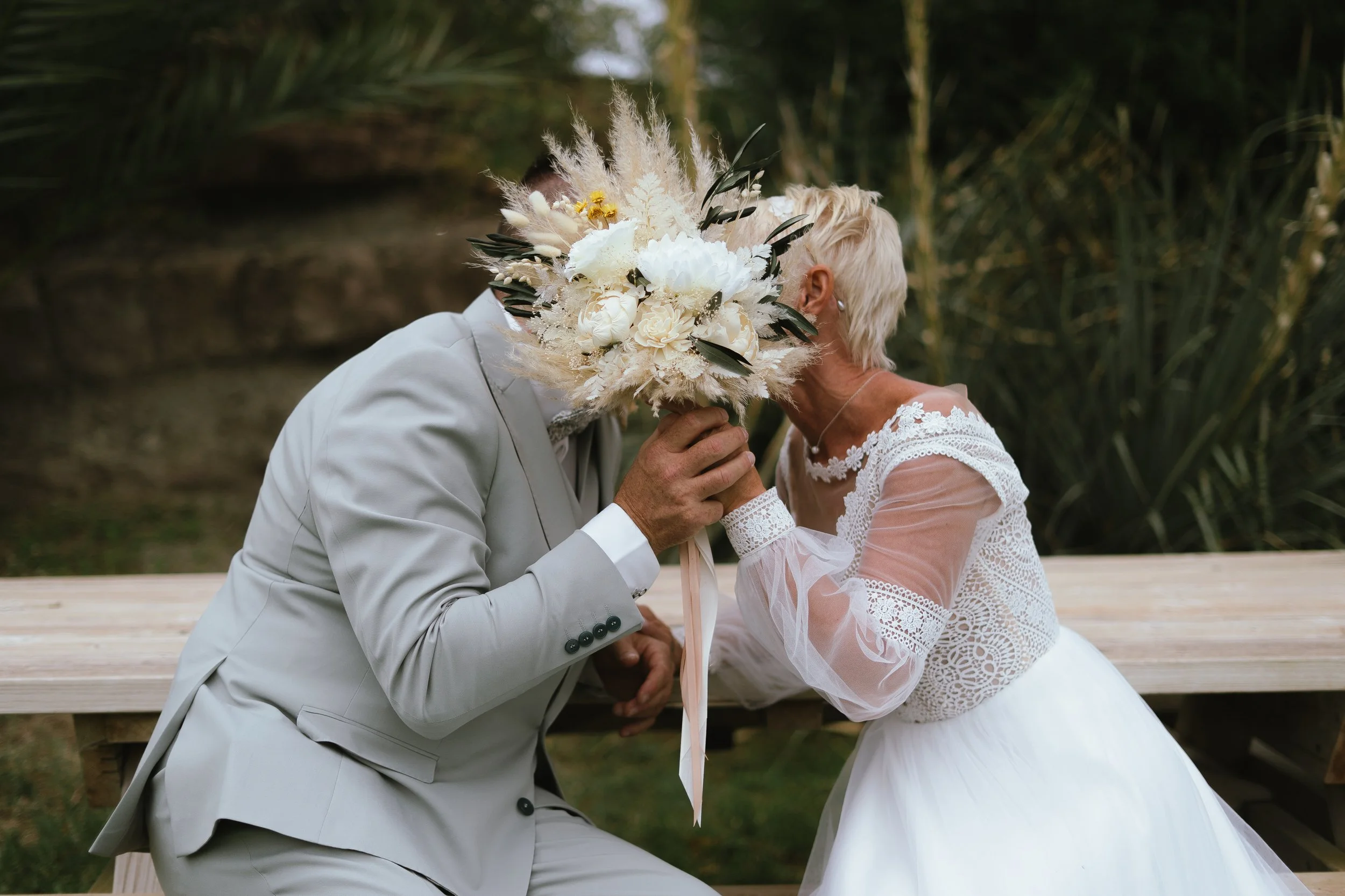 Un couple de mariés échangeant un baiser avec un bouquet de fleurs devant un fond naturel.