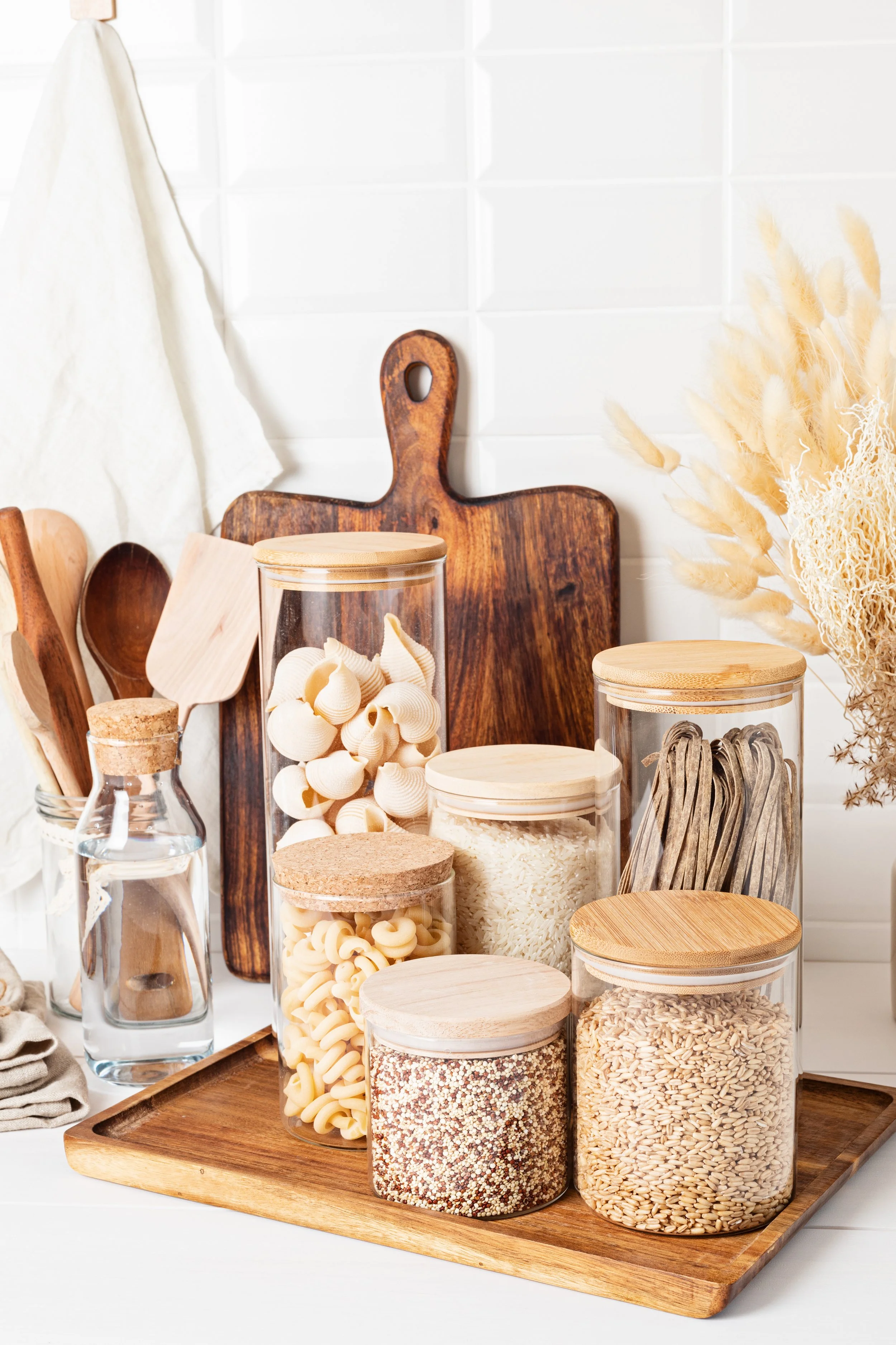 Kitchen storage jars filled with pasta, rice, and grains on a wooden tray.