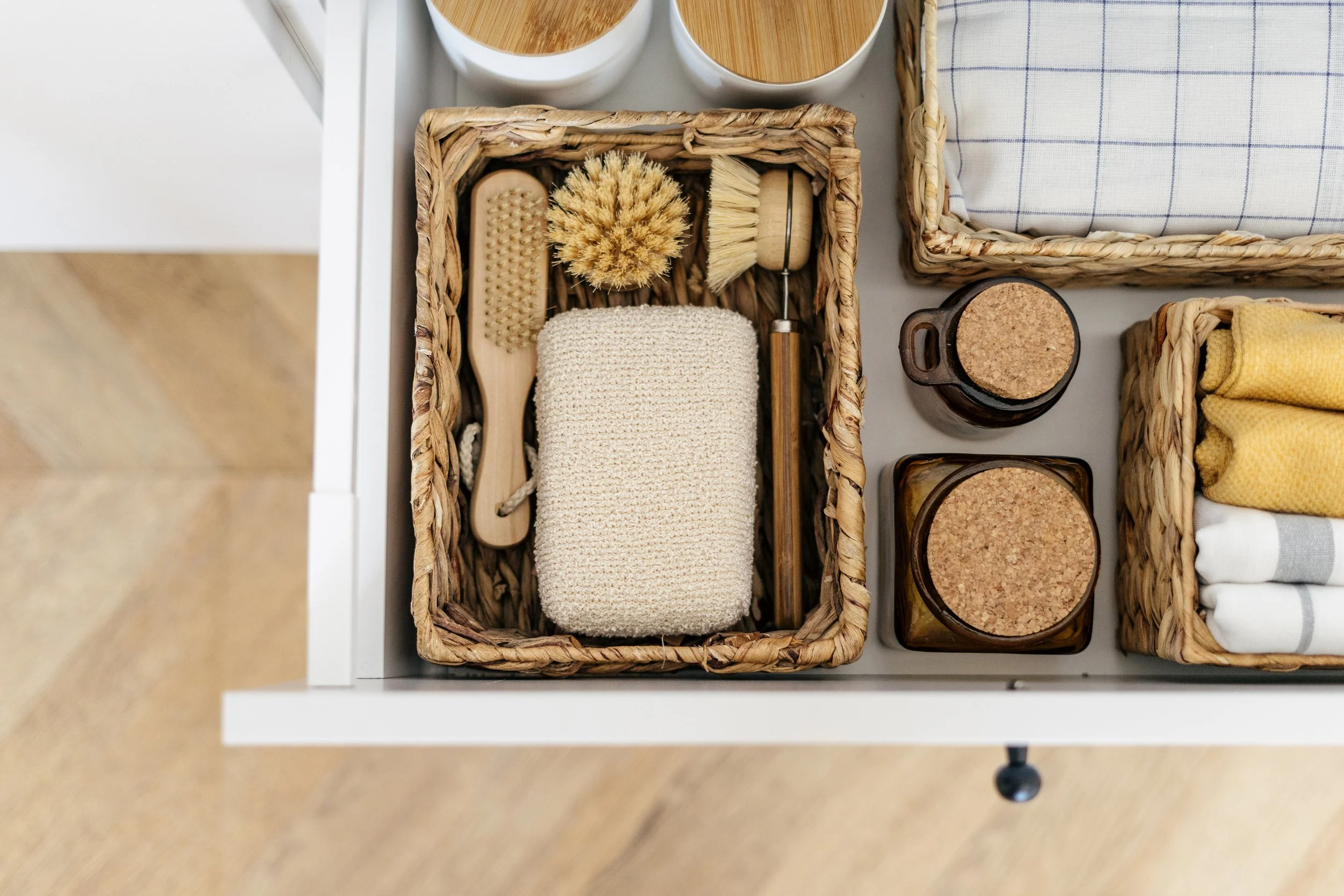 A drawer containing natural fiber baskets with personal care items. One basket holds a wooden body brush, a scrubbing brush, a loofah, and a soap dish. Adjacent are jars with cork lids and cloth towels, organized in different compartments.