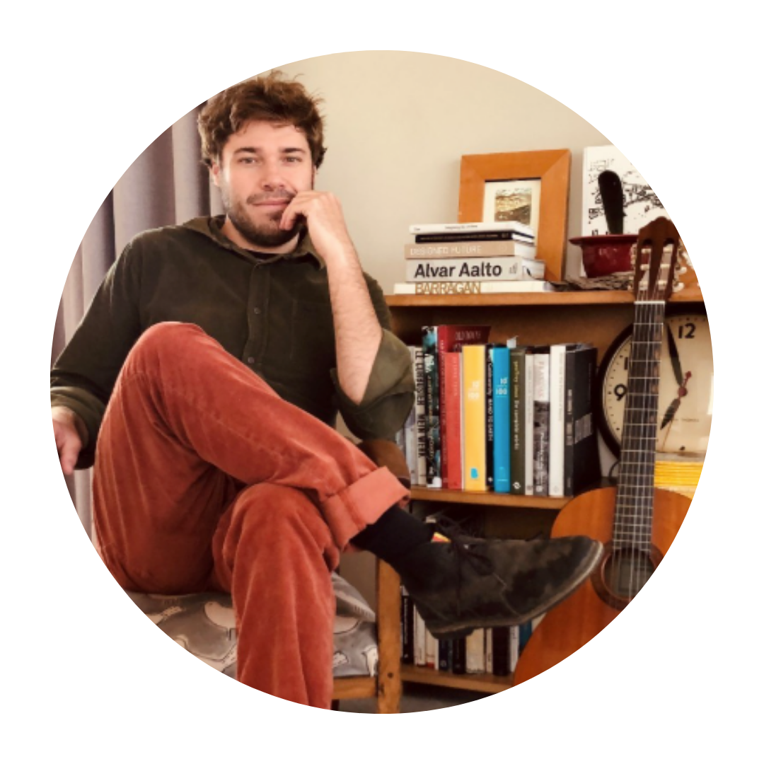 A young man with curly hair and a beard sitting on a chair in a room, with a bookshelf, a guitar, and a clock behind him.