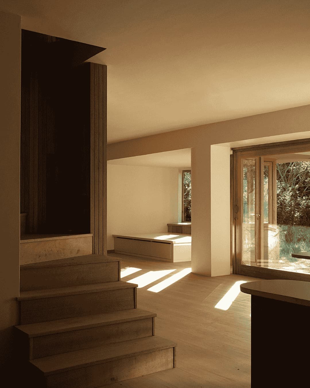 Interior of a modern, minimalist home with wooden floors, steps, and large windows letting in natural sunlight.