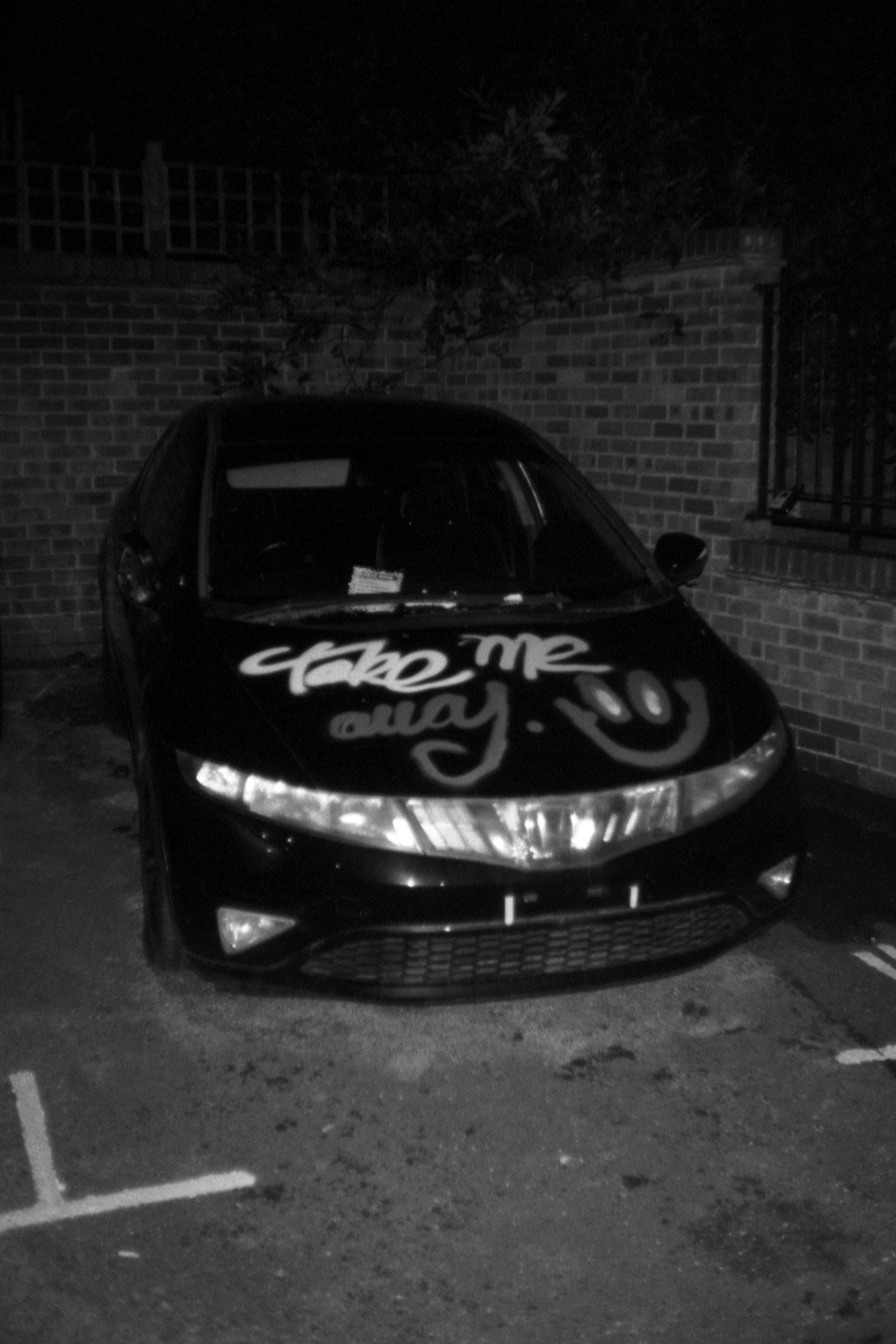 “Black-and-white photo of a parked car at night with ‘take me away’ and a smiley face painted on the hood, evoking the longing to rewrite or escape personal memories.