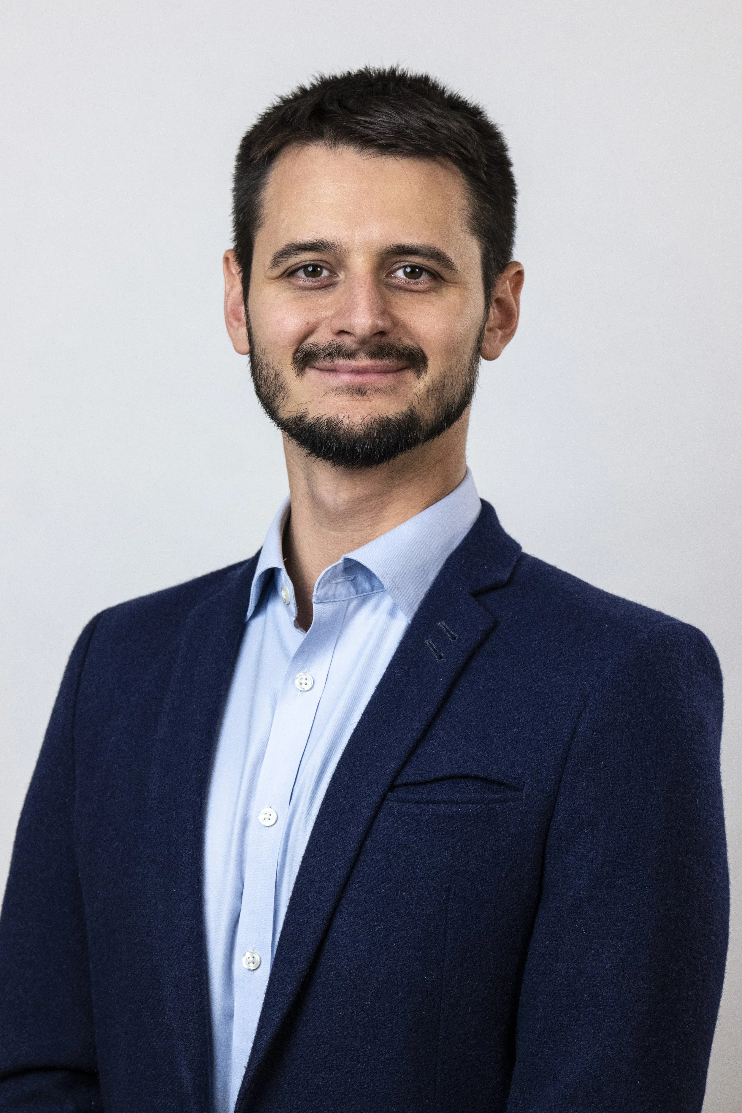 A man with dark hair and a beard wearing a blue blazer and light blue shirt, smiling at the camera against a neutral background.