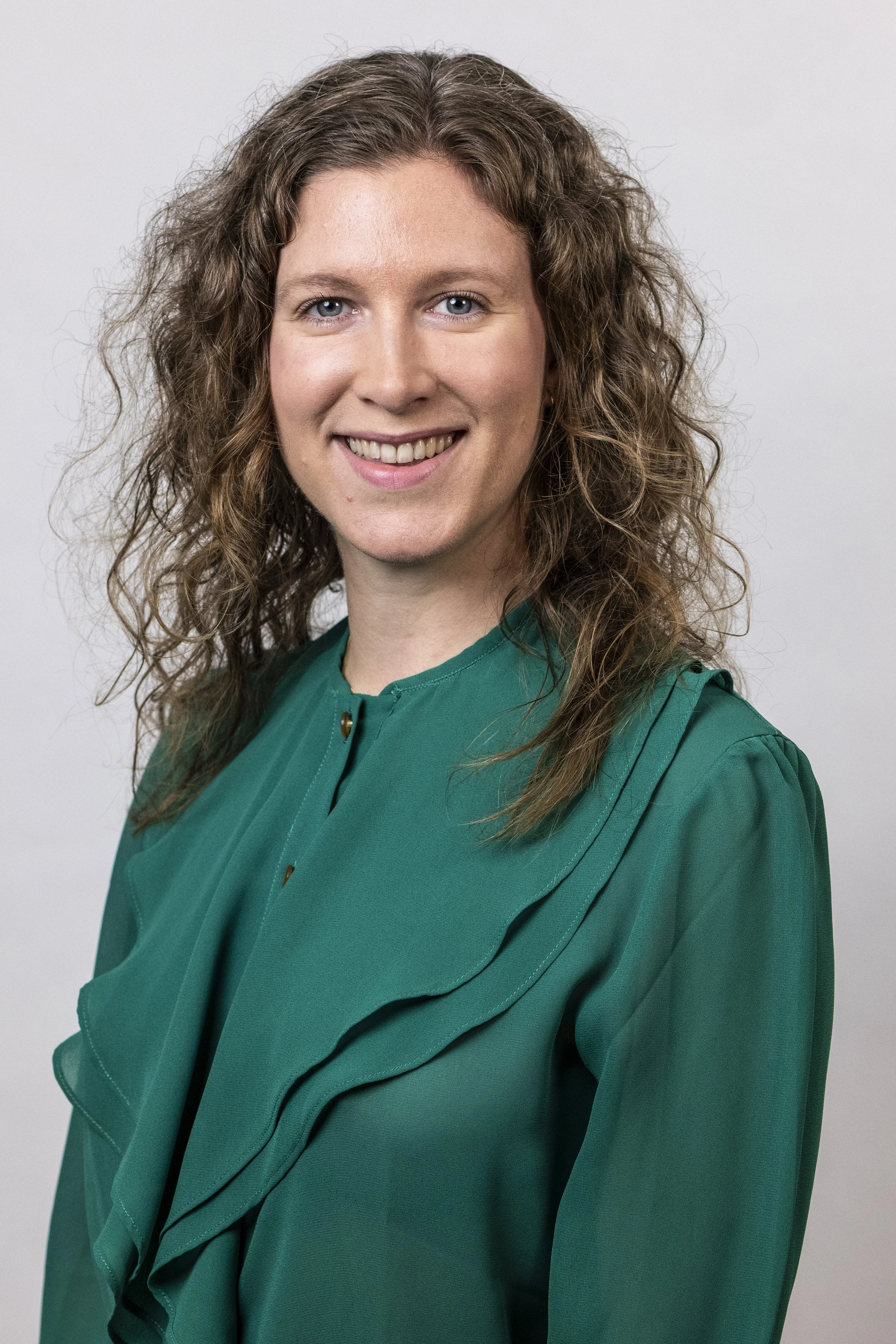 A woman with wavy, shoulder-length blonde hair and blue eyes, smiling, wearing a green blouse, against a neutral background.