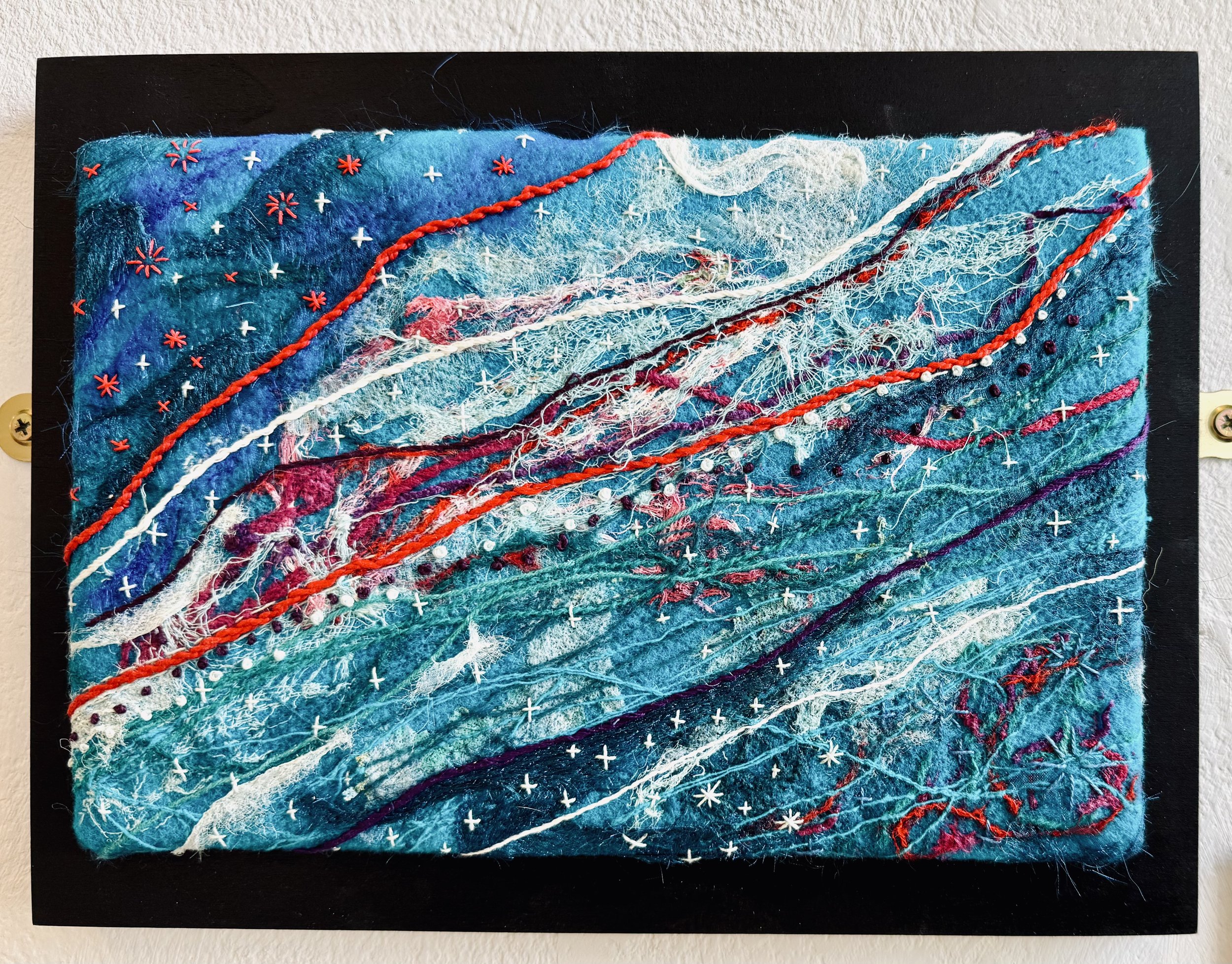Textile art on a black background with various shades of blue, white, red, and purple threads and embroidery, resembling a stylized abstract landscape or ocean scene.