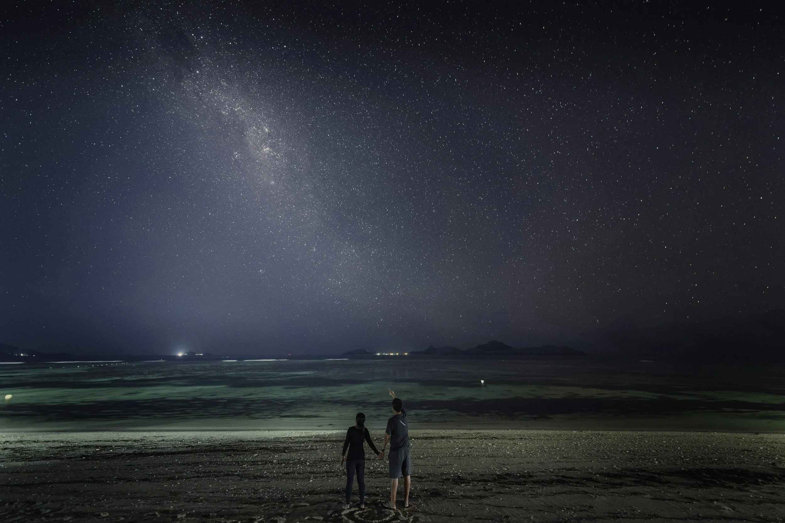 A couple stand together by the sea beneath the Milky Way in a star-filled night sky