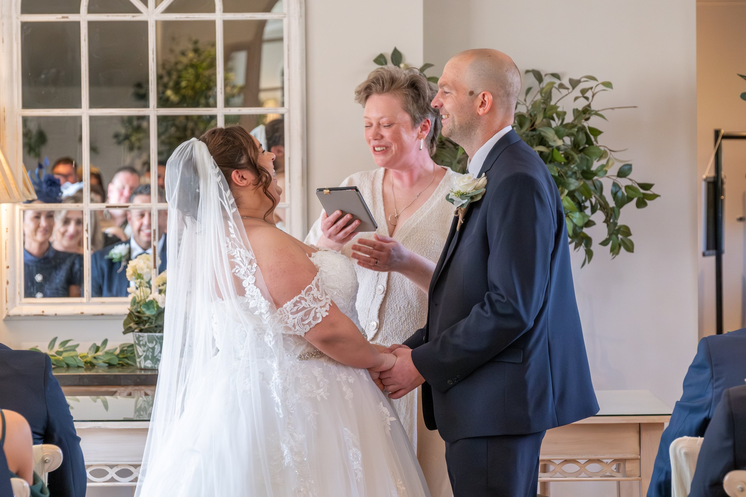 A bride and groom holding hands during a wedding ceremony officiated by a woman with a tablet, with a mirror reflecting guests in the background.