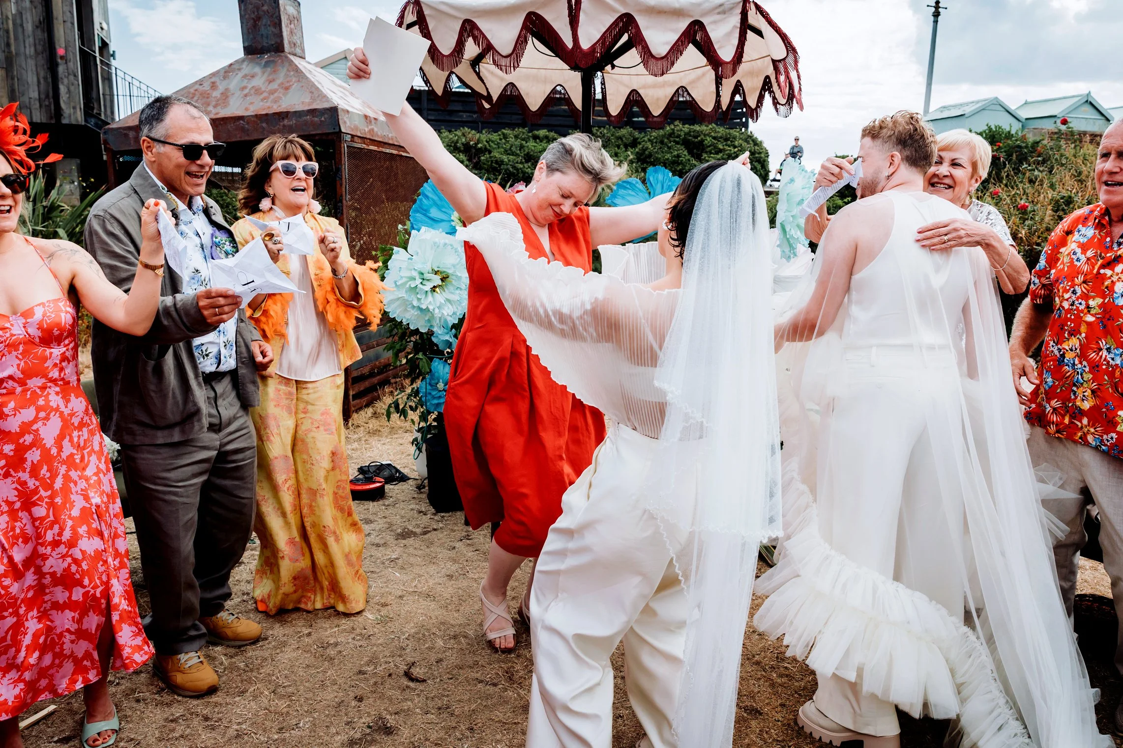 A celebrant goes to embrace the couple surrounded by family and friends at a colourful outdoor wedding ceremony.