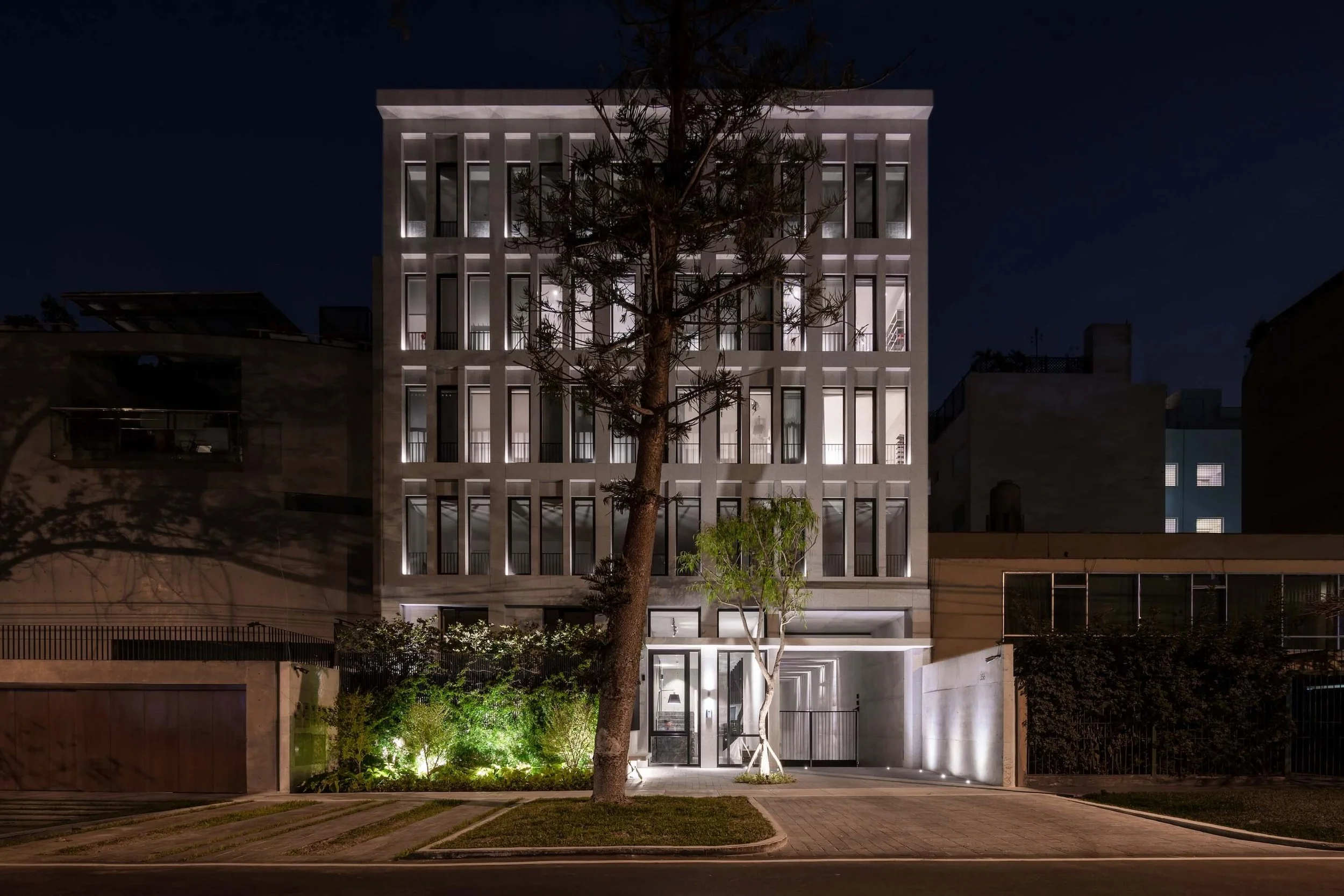 Edificio moderno de noche con fachada de ventanas y luces en la noche, rodeado de árboles y vegetación en el frente.