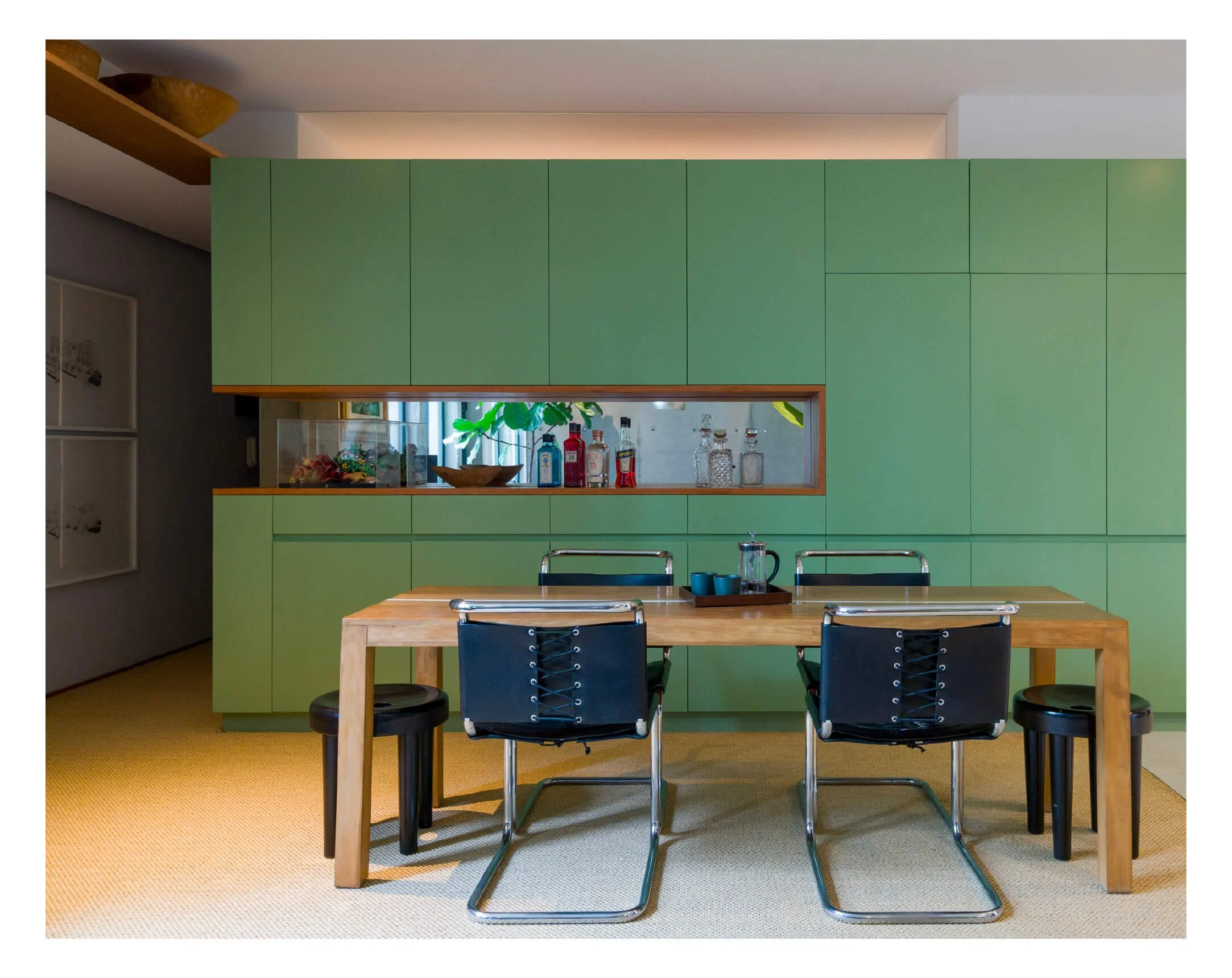 Sala de comedor moderna con mesa de madera y cuatro sillas negras con estructura metálica, fondo con armario verde y ventana decorativa con plantas y botellas decorativas.