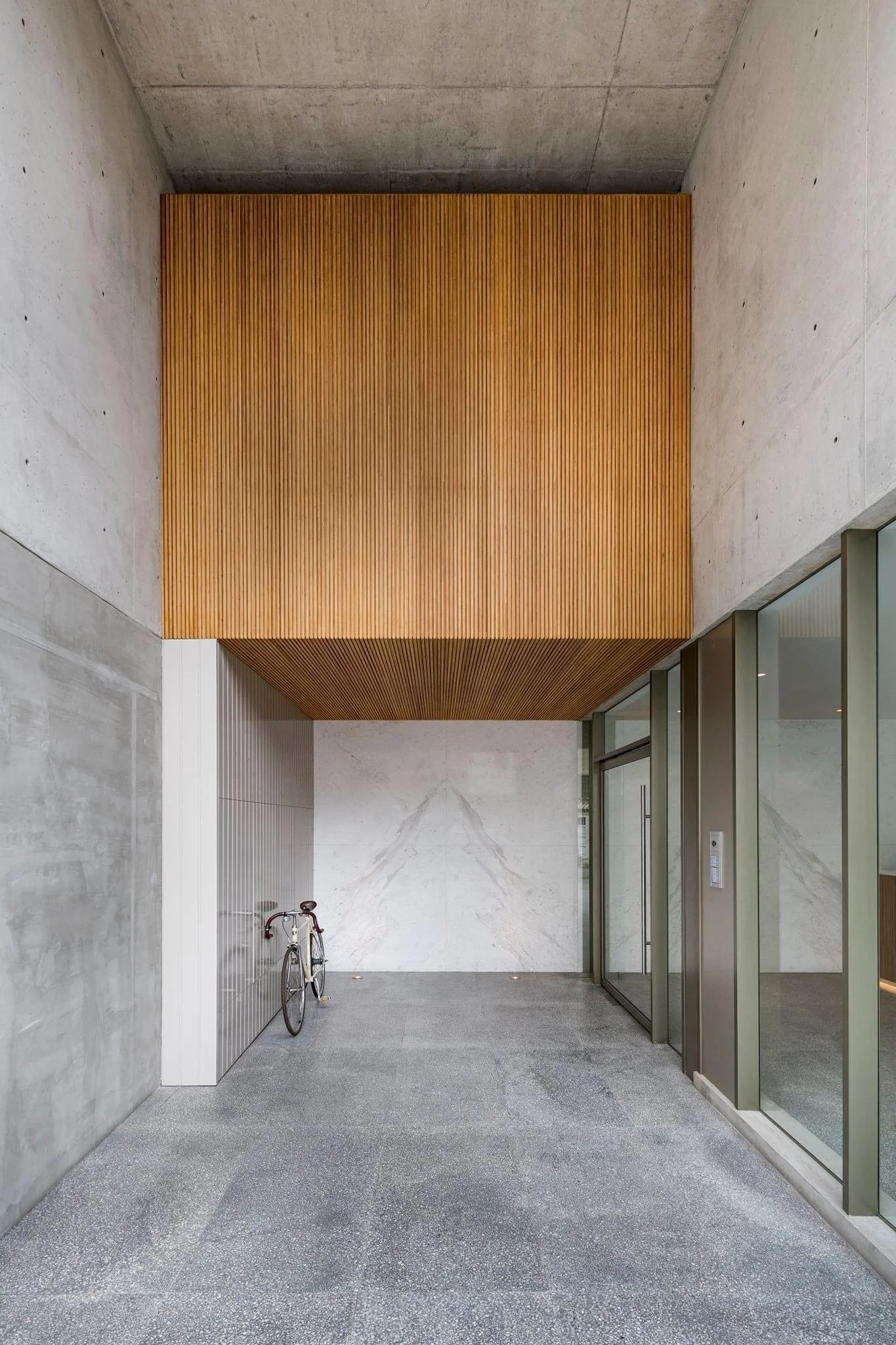 Entrada de edificio moderna con paredes de concreto, borde de madera suspendido y vestíbulo con suelo de granito. Hay una bicicleta apoyada en la pared y puertas de cristal a la derecha.