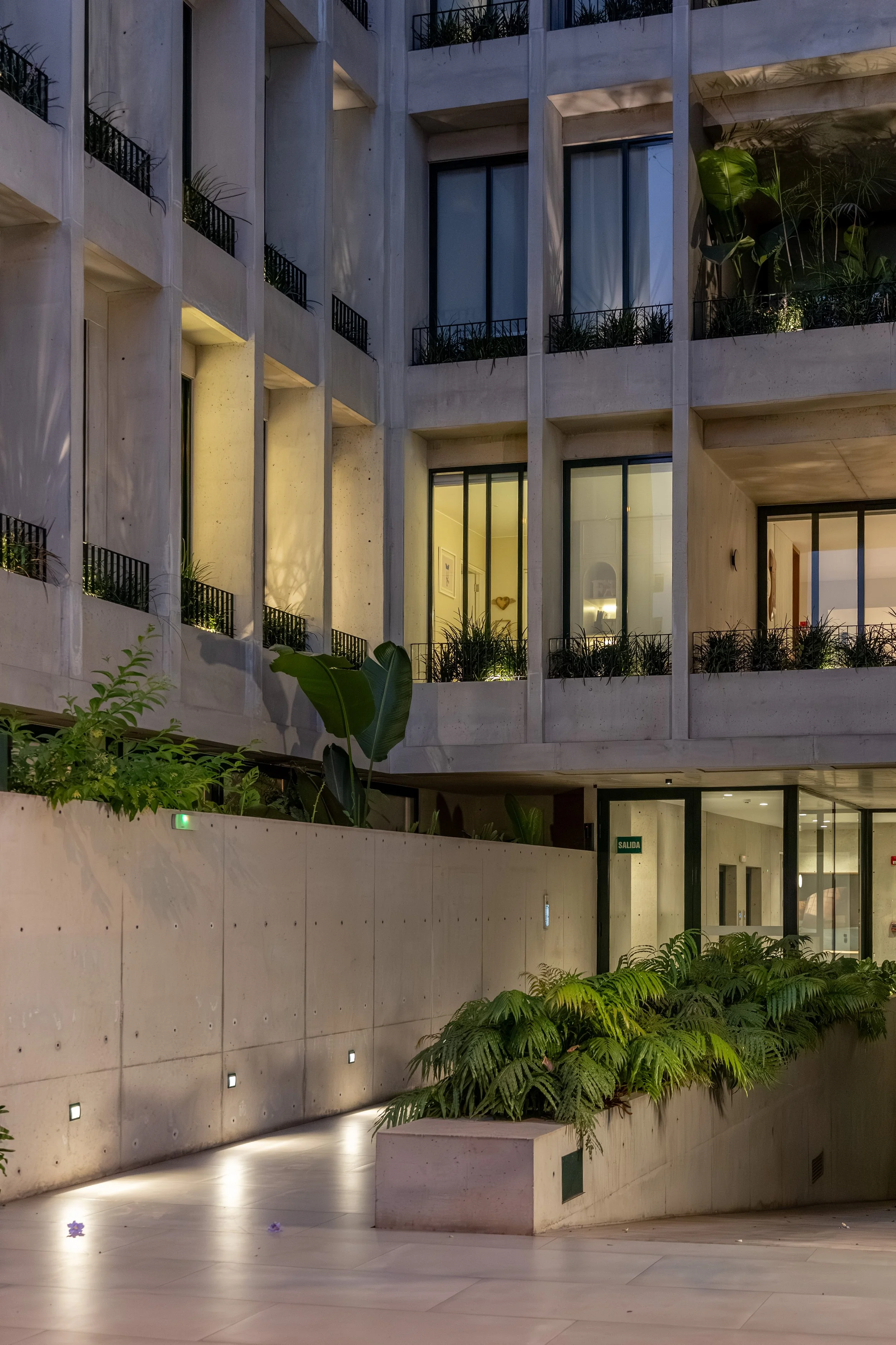 Imagen de un edificio moderno de varios pisos con balcones y ventanas de vidrio, iluminado con luz interior y ambiental por la noche, con áreas con plantas verdes en el exterior.