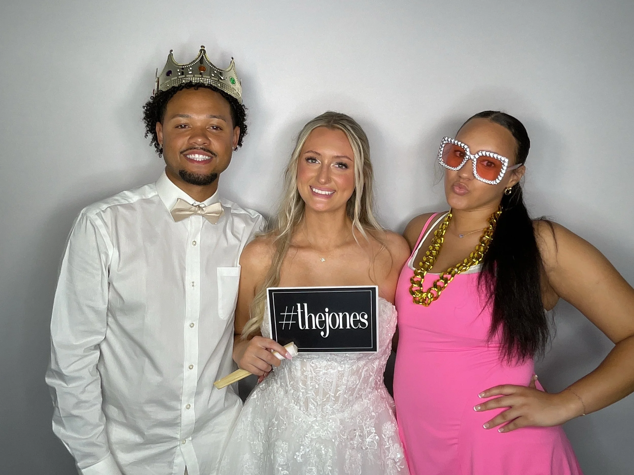 Three people at a celebration event, one man wearing a crown and bow tie, woman in a white dress holding a sign, woman in a pink dress wearing large jewelry and sunglasses.