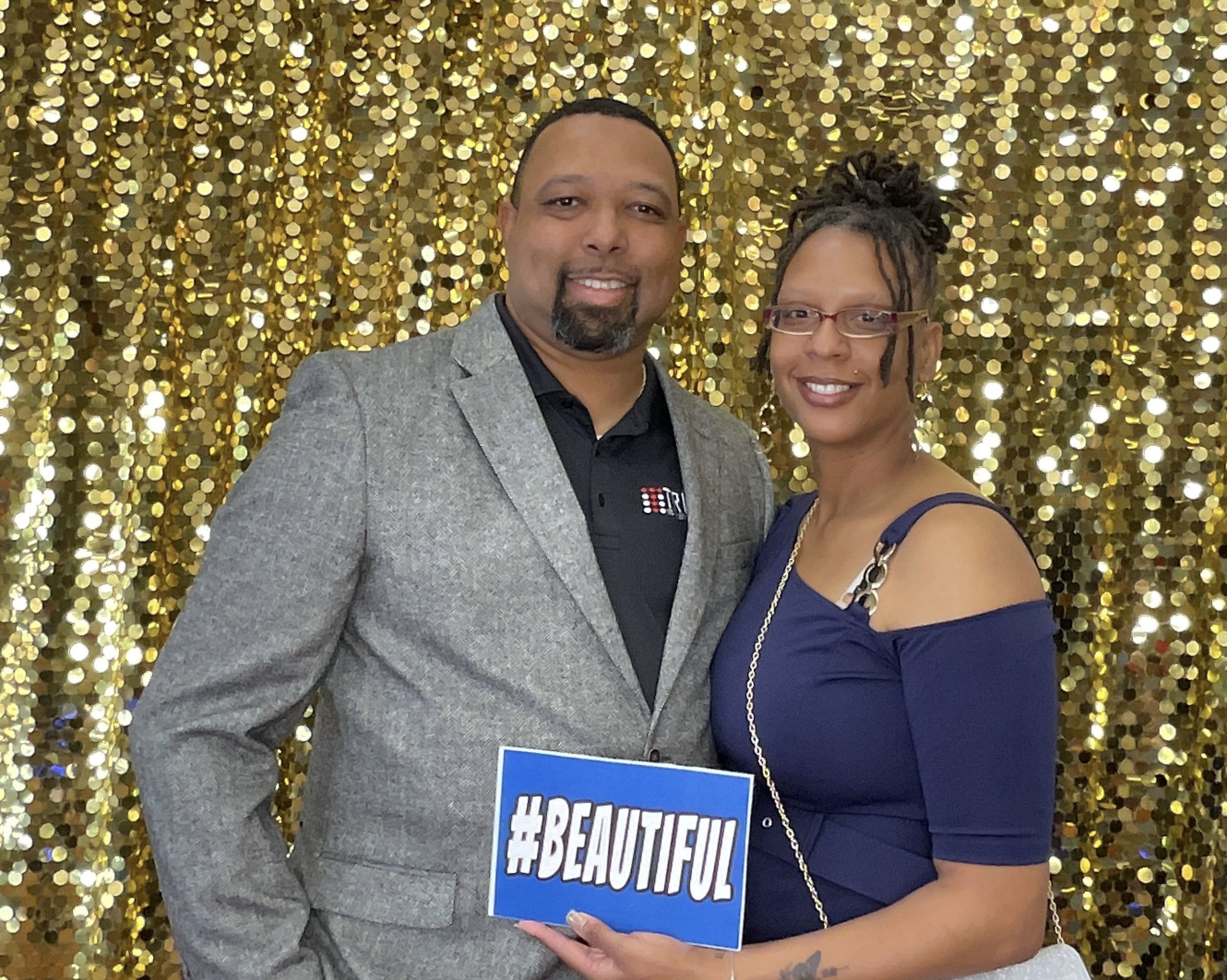 A man and woman smiling in front of a gold sequin backdrop, holding a sign that says '#BEAUTIFUL'. The man is wearing a gray suit jacket, and the woman is in a dark blue off-shoulder dress.