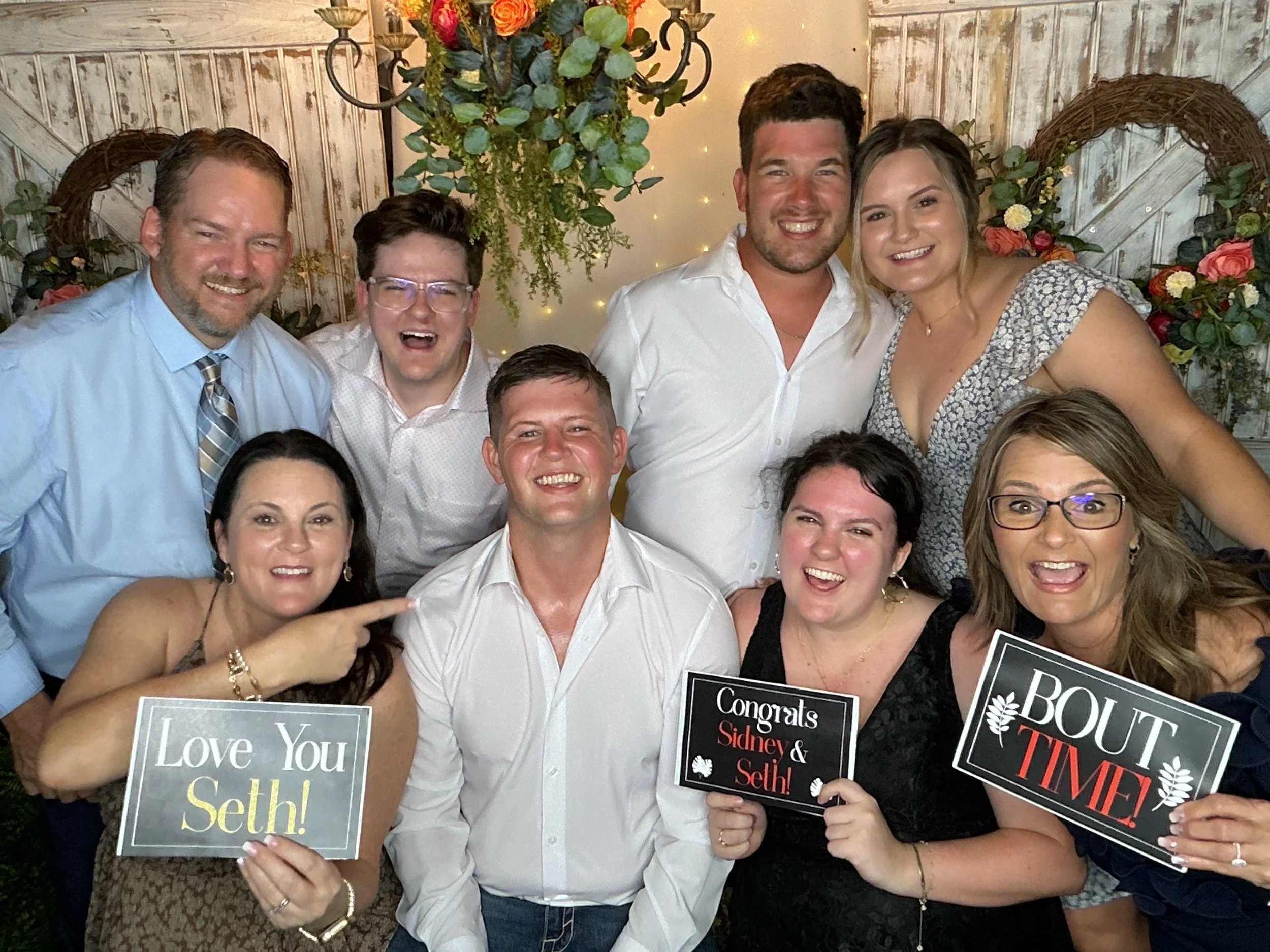 A group of people celebrating at a wedding reception, holding signs that say 'Love You,' 'Congrats Sidney & Seth,' and 'BOUGHT TIME.' The backdrop features floral arrangements and warm lighting.