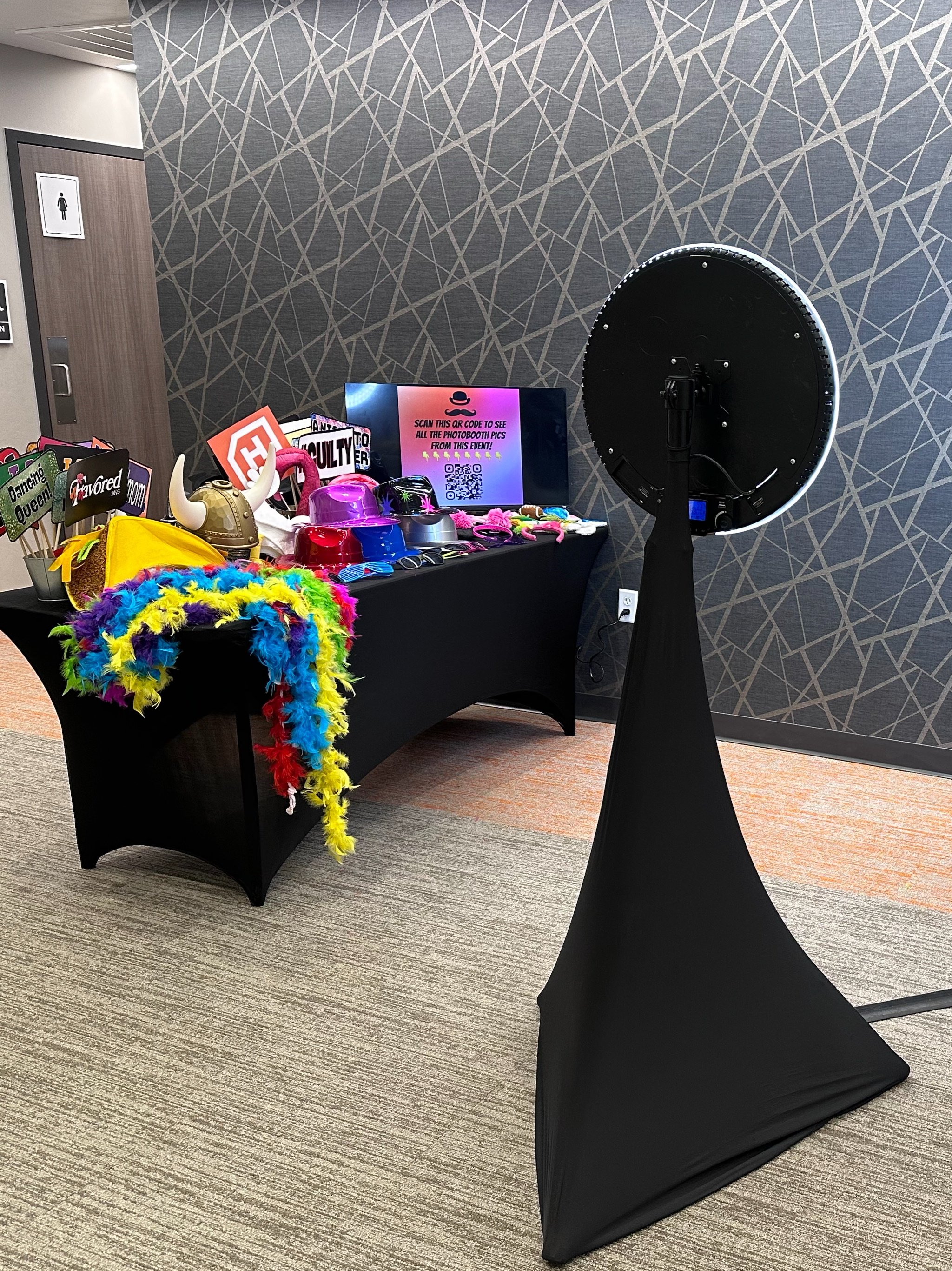 Photo booth setup with a ring light on a stand, surrounded by tables with colorful props including hats, signs, and feather boas.