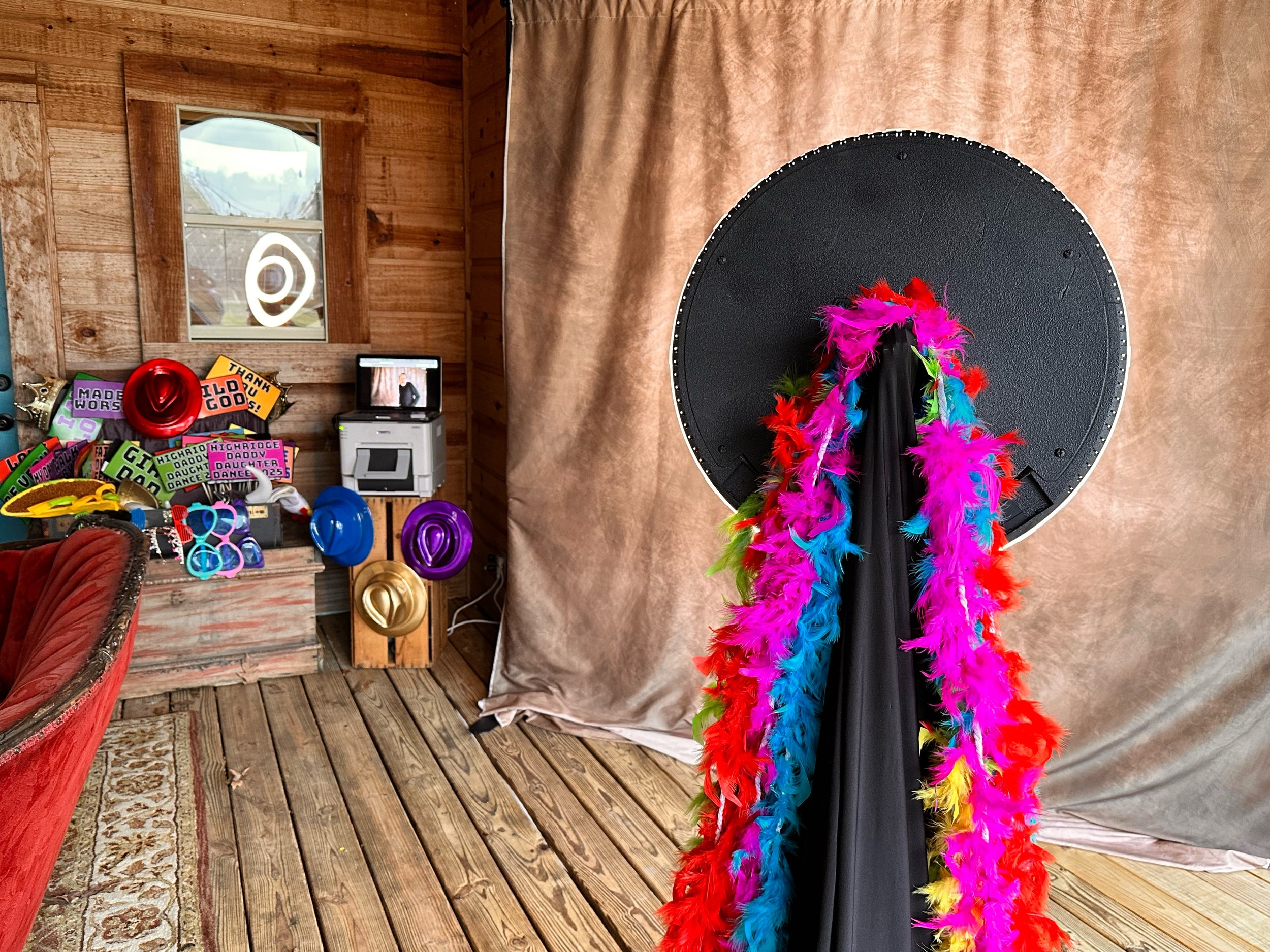 Photo booth setup with colorful feather boas and props, including hats and signs, in a rustic wooden room with a window and curtain backdrop.