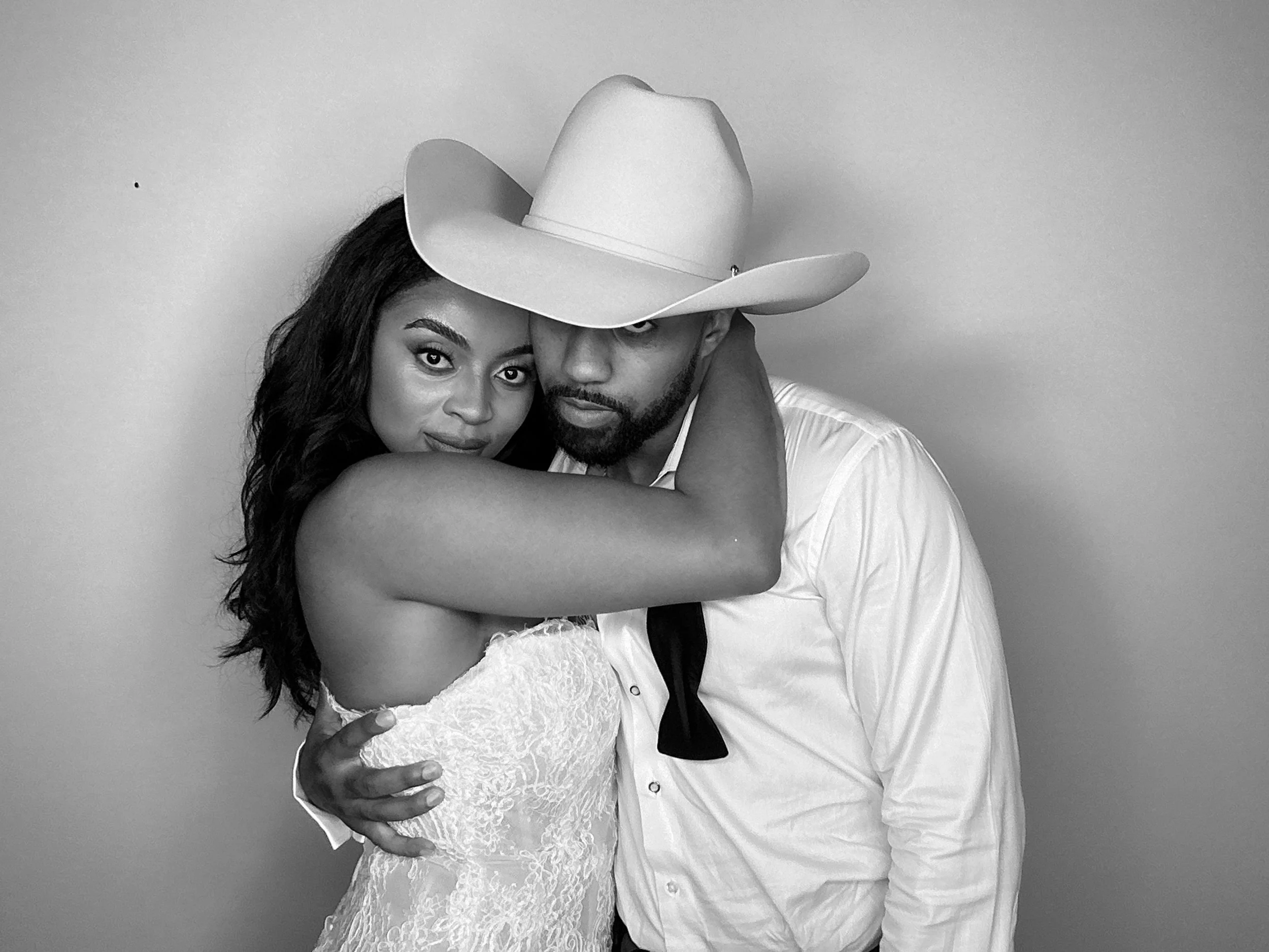 A black and white photo of a couple embracing, with the woman wearing a lace dress and the man wearing a white shirt and a cowboy hat.