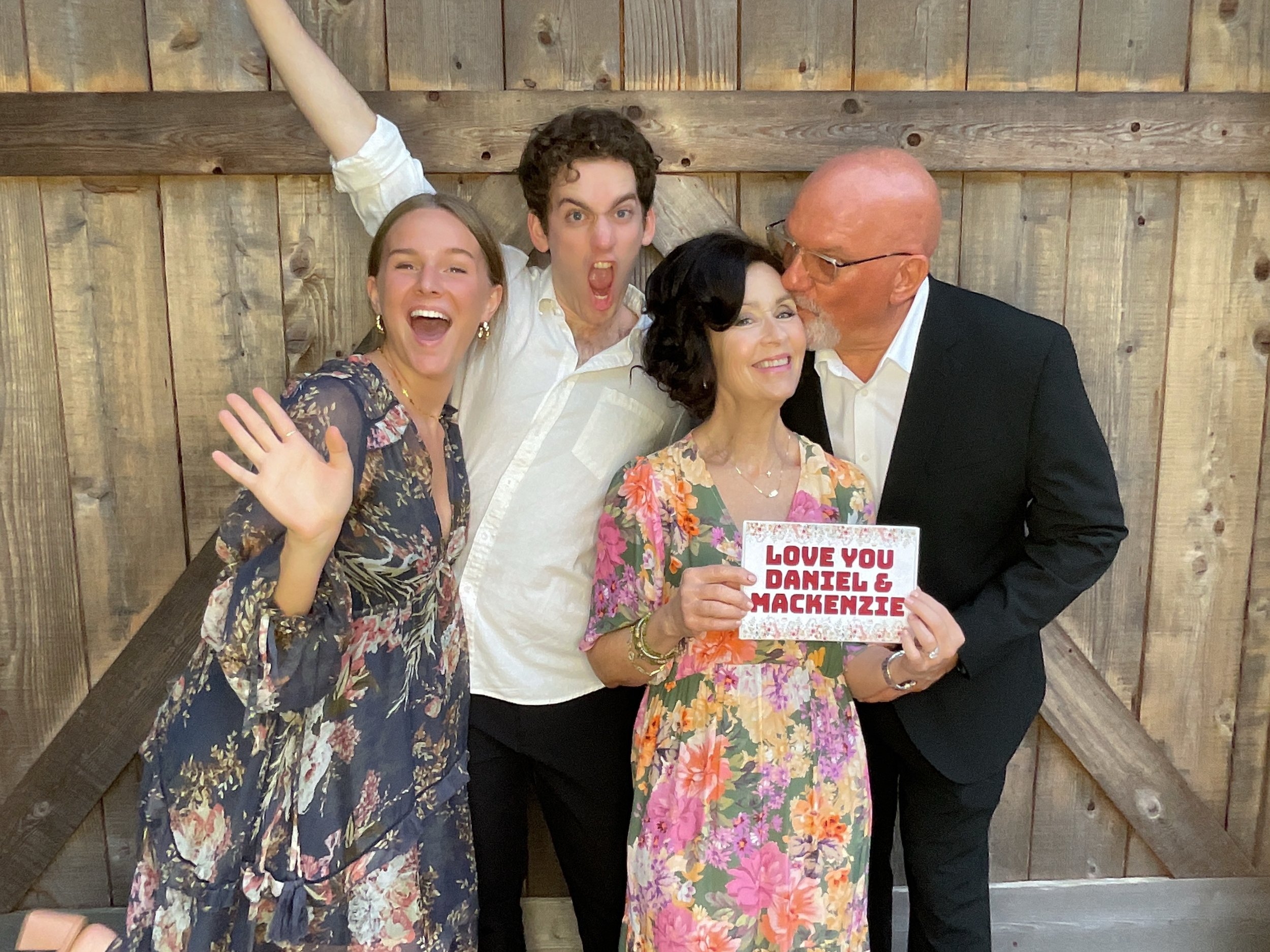 Four people posing cheerfully in front of a wooden wall, with one person holding a sign that reads 'Love You Daniel & Mackenzie.' Two are wearing floral dresses, one in a white shirt, and the other in a suit.