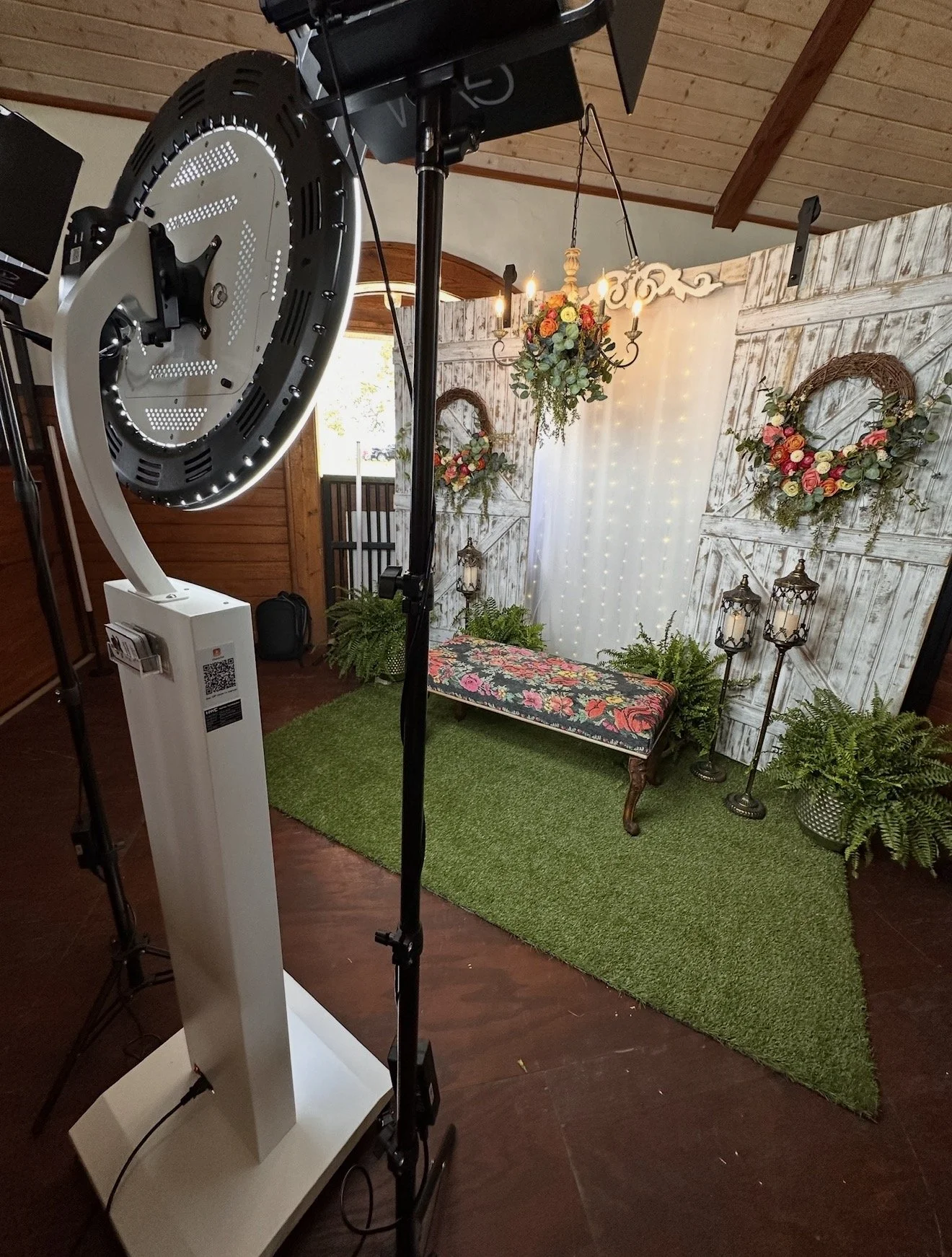 A decorated photo booth setup with a floral backdrop, wreaths, lanterns, potted ferns, a floral cushion bench, and twinkling string lights.