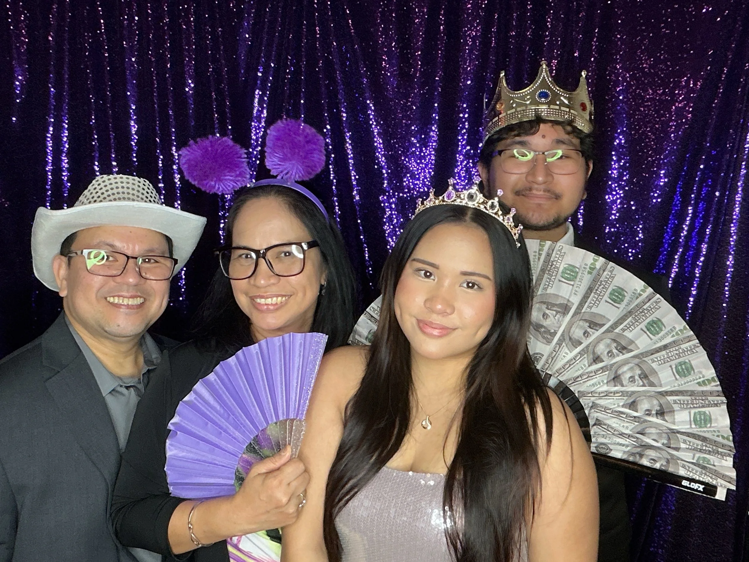 A group of five people posing for a photo at a celebration or party. Two women wearing tiaras. One woman holding a purple paper fan, another with a necklace. One man wearing a crown and glasses, with dollar bills on his shoulder. The other man has a white hat and glasses. They are against a backdrop of shiny purple and black curtains with purple pom-poms.