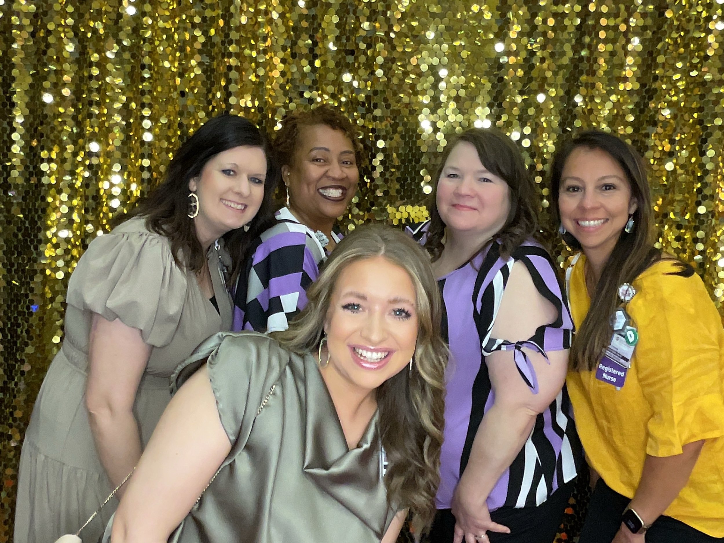 Group of five women smiling in front of a gold sequin backdrop.