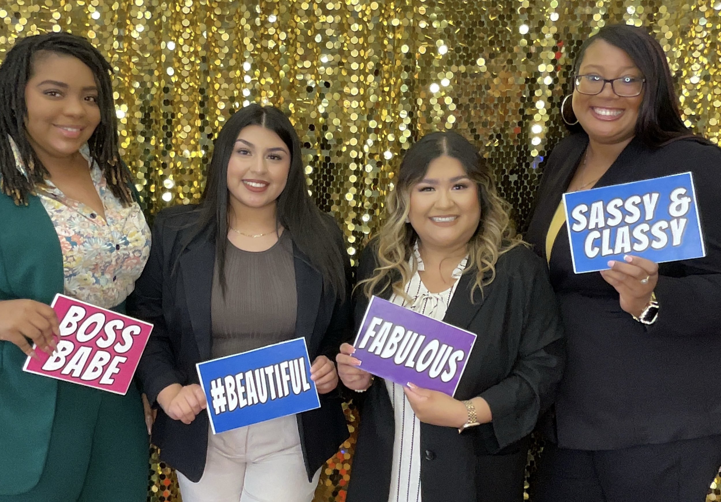 Four women holding signs with positive affirmations against a gold sequin backdrop.