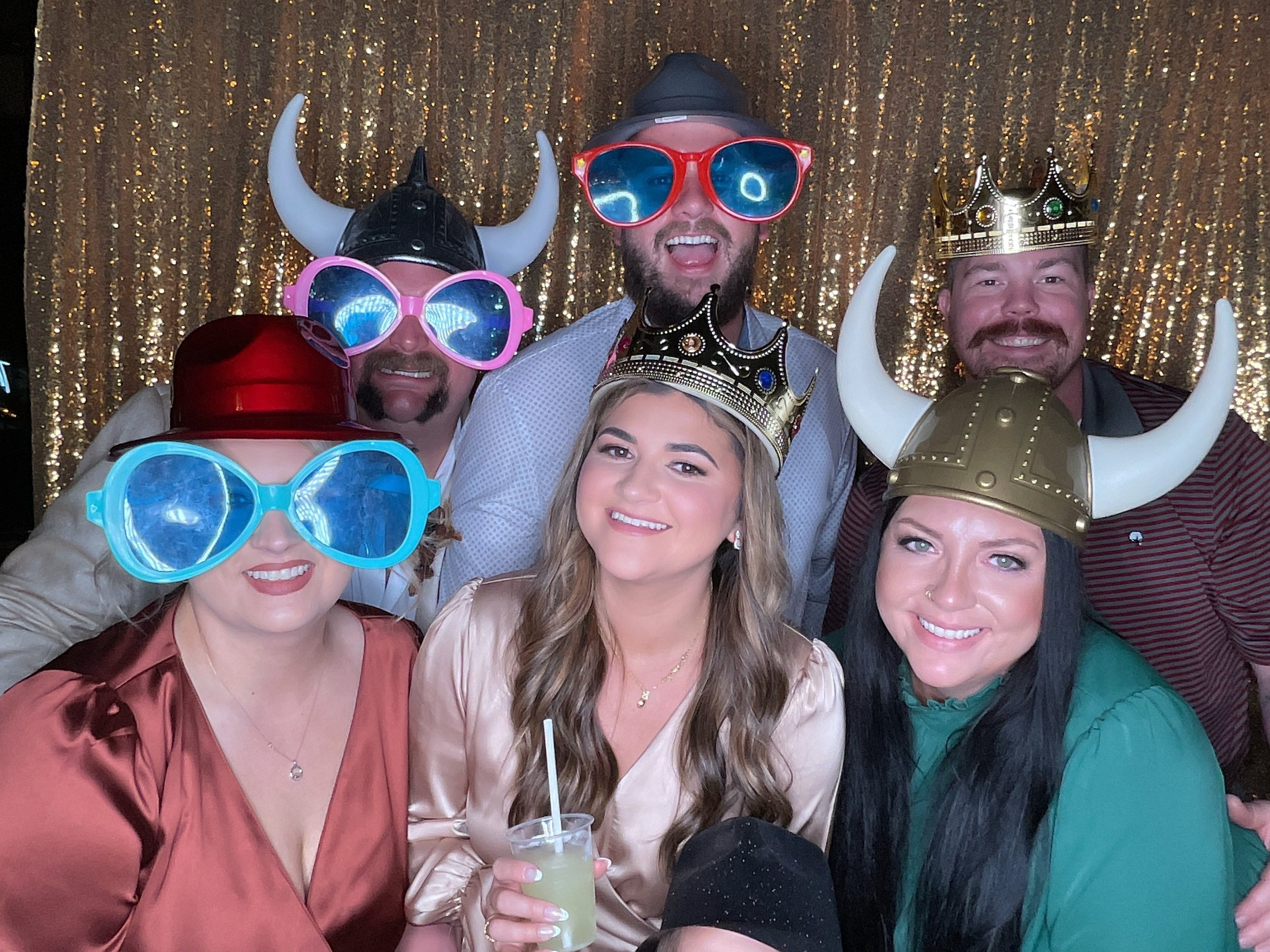 Group of people wearing novelty hats, oversized glasses, and crowns in front of a golden sequin backdrop, smiling at the camera.
