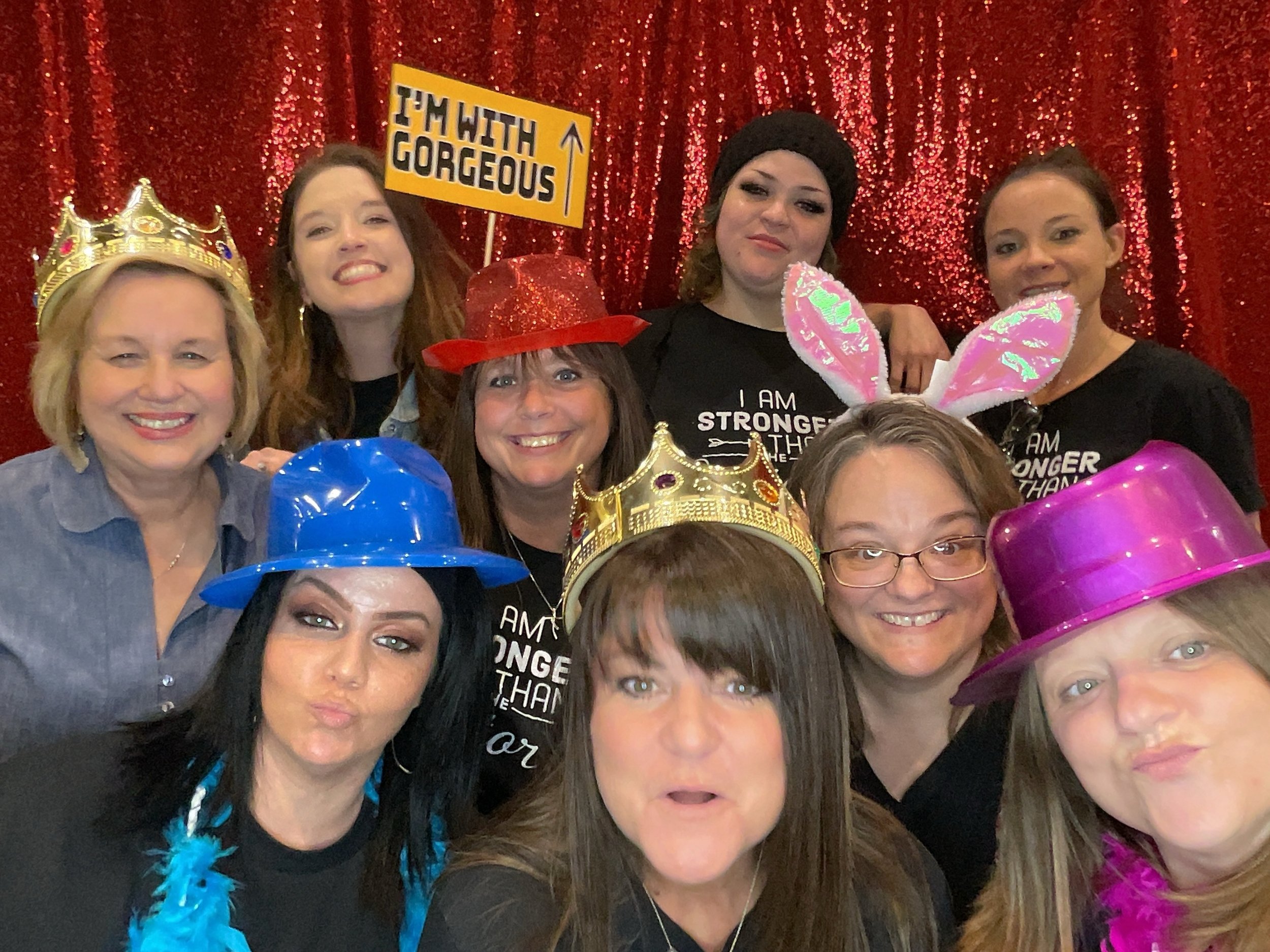 A group of eight women wearing various novelty hats and accessories in front of a red sequined backdrop. They are posing for a fun photo, with one holding a sign that says "I'm with gorgeous."