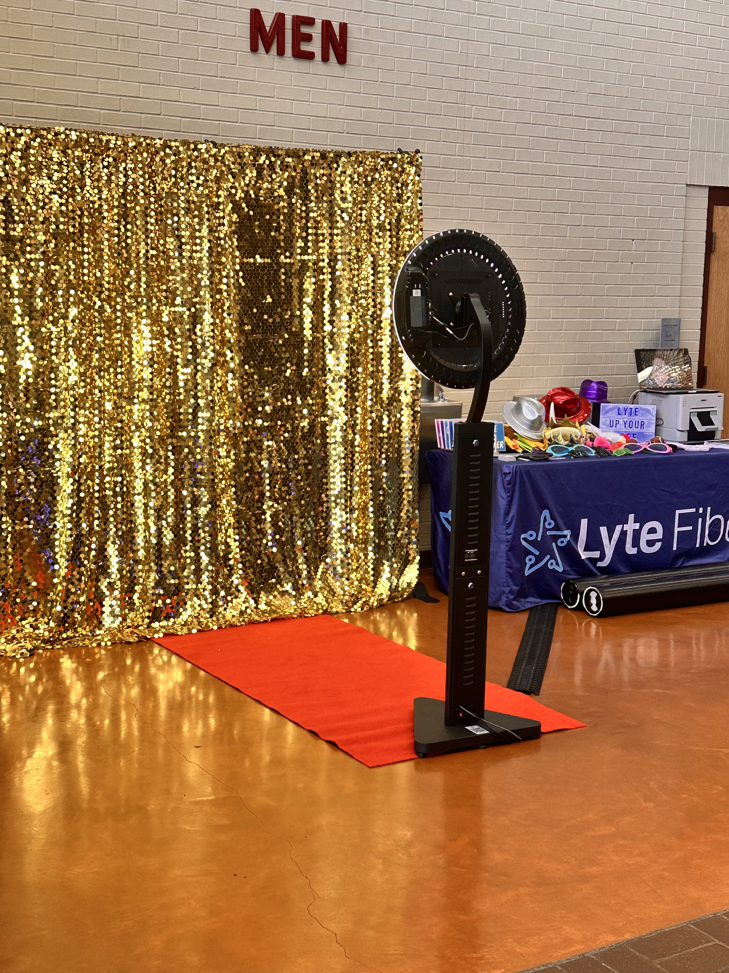 Photo booth setup with a gold sequin backdrop, red carpet, and a ring light on a stand, next to a table with props and a 'Lyte Fiber' banner.