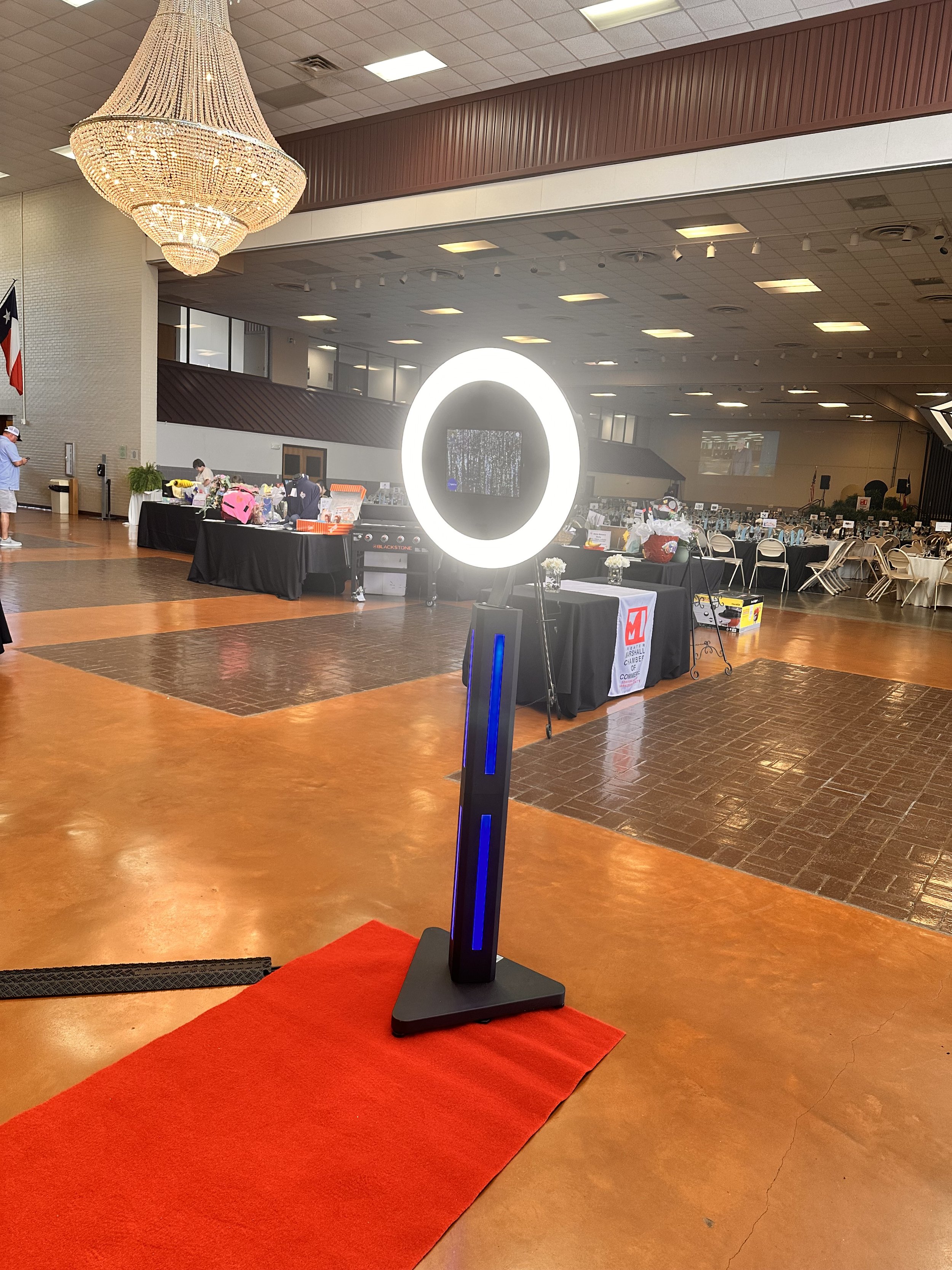 Indoor event space with chandelier, photo booth ring light, red carpet, and tables with various items.