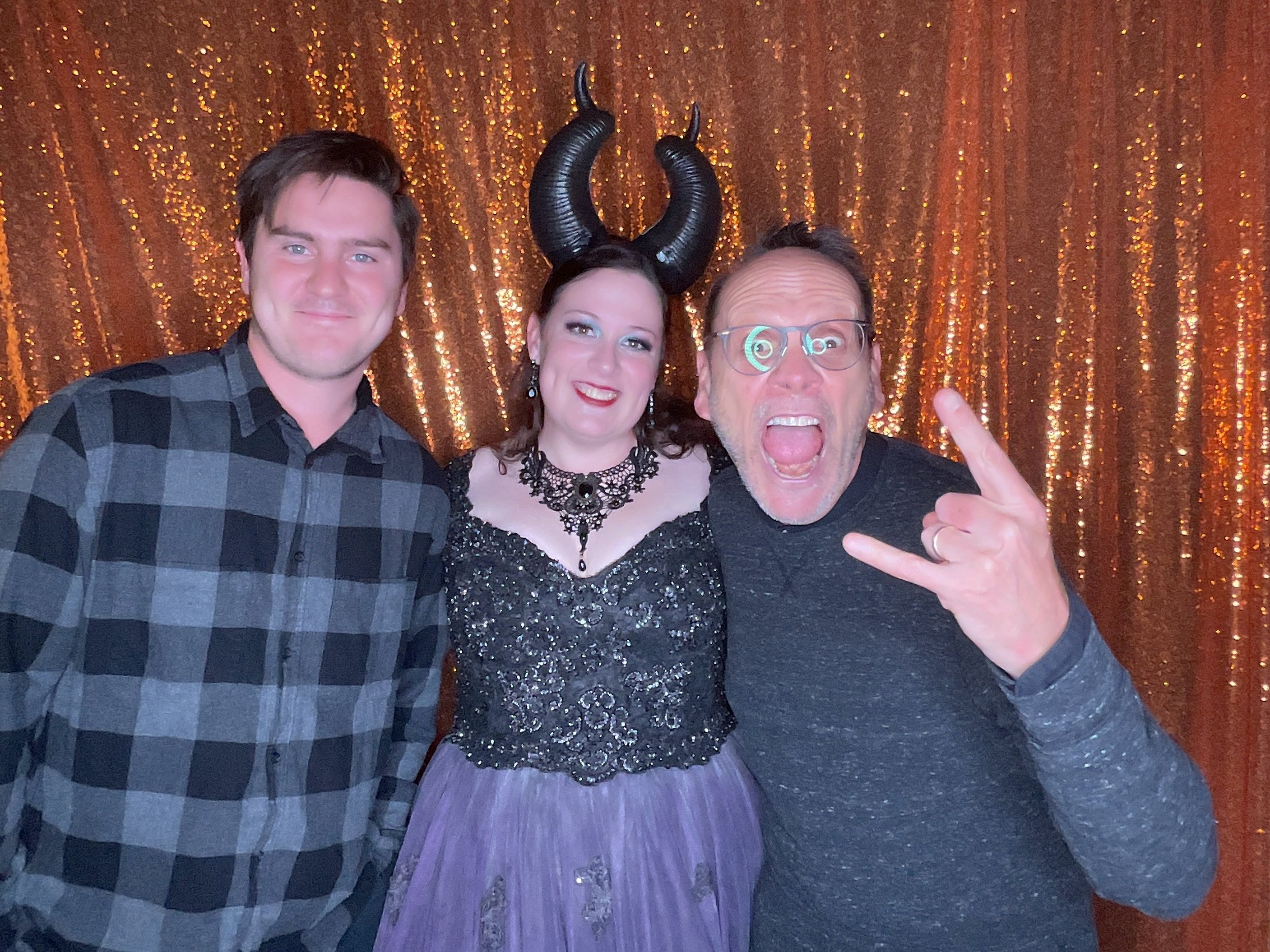 Three people in front of a glittery gold backdrop. One person wears a black and purple costume with horned headpiece, flanked by two others, one in a plaid shirt, the other wearing glasses making a hand gesture.