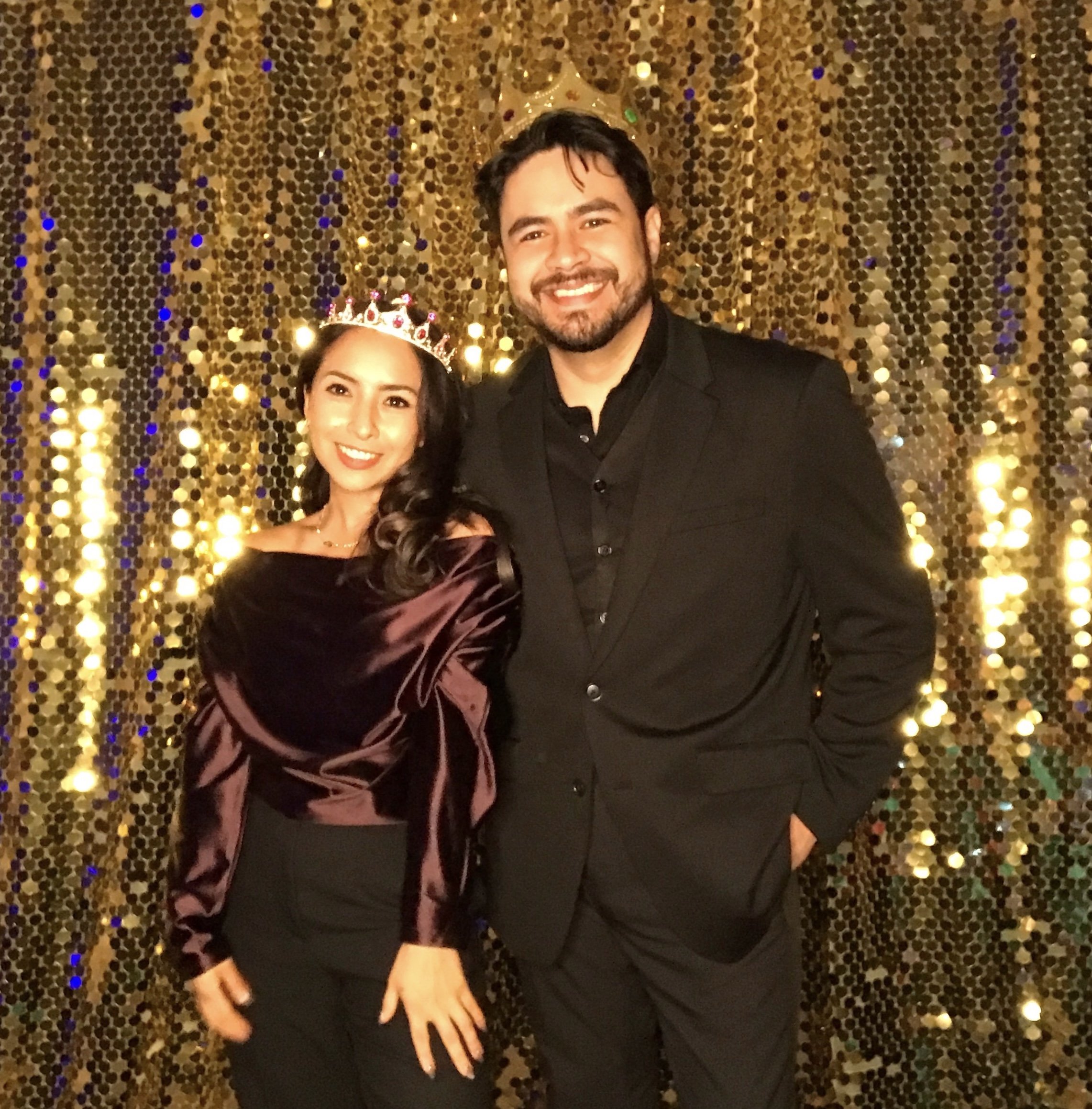 A man and woman dressed in formal attire wearing crowns, posing in front of a gold sequin backdrop.
