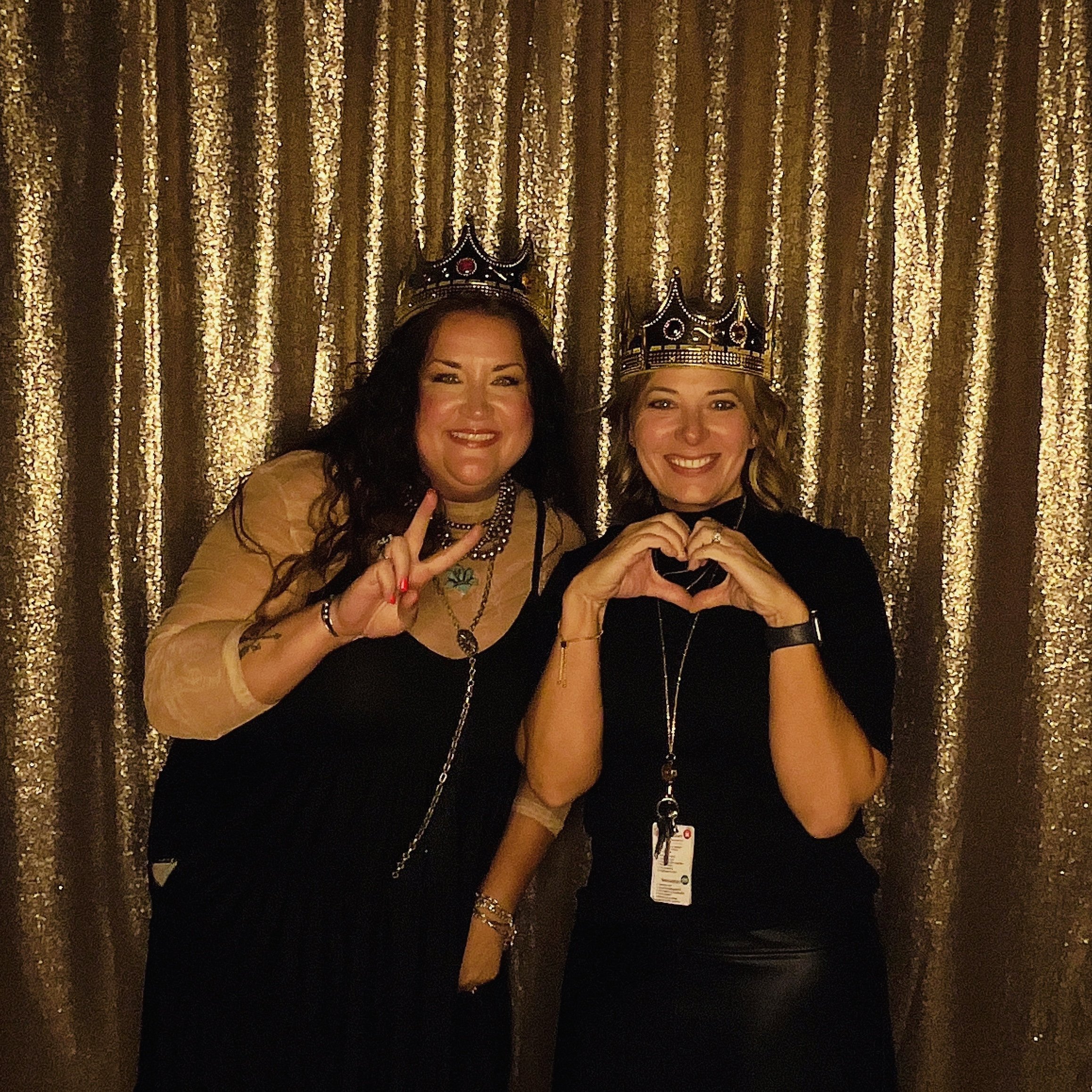 Two women wearing crowns in front of a gold shimmering curtain, one making a peace sign and the other forming a heart with their hands.