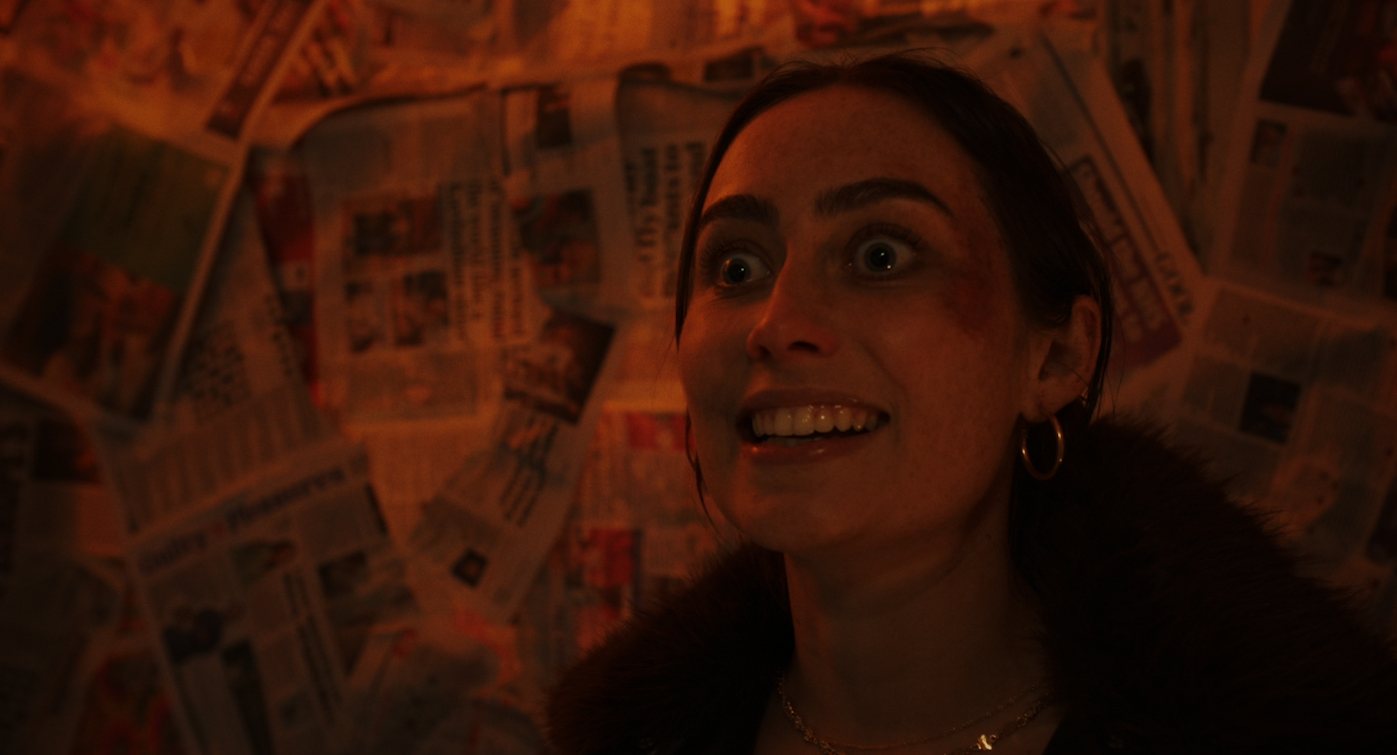 A close-up of a woman with dark hair, wearing gold hoop earrings and a fur coat, smiling with bright eyes, standing against a wall covered in newspapers.