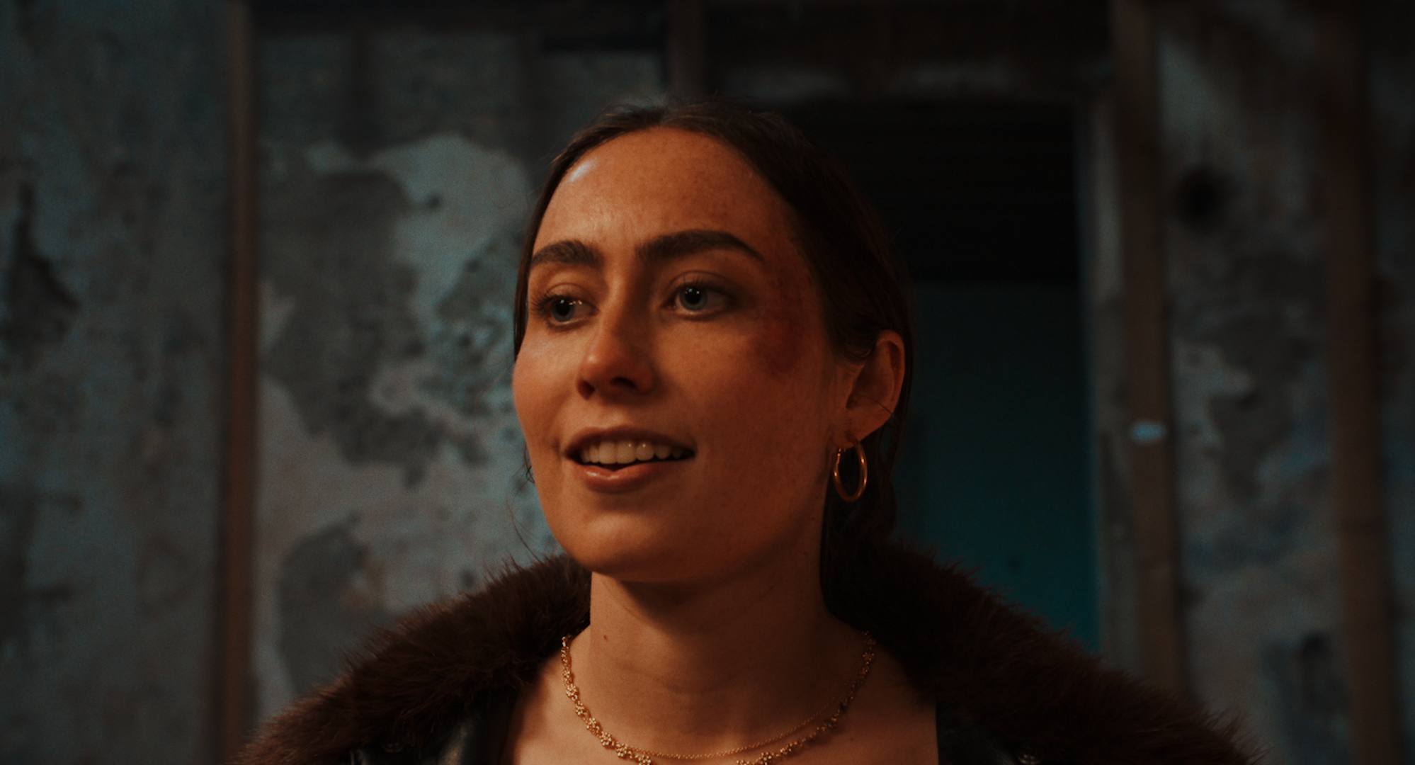 A woman with brown hair, blue eyes, and freckles, wearing hoop earrings and a gold necklace, smiling slightly. She is dressed in a dark fur coat and is in front of a textured, grayish rock wall.