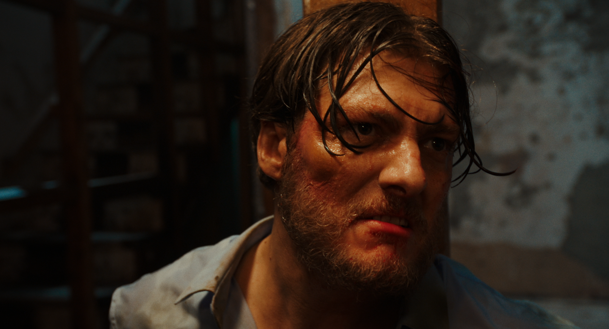 Close-up of a man with wet hair and weathered face, looking intently to the side, with a dark, smoky background.