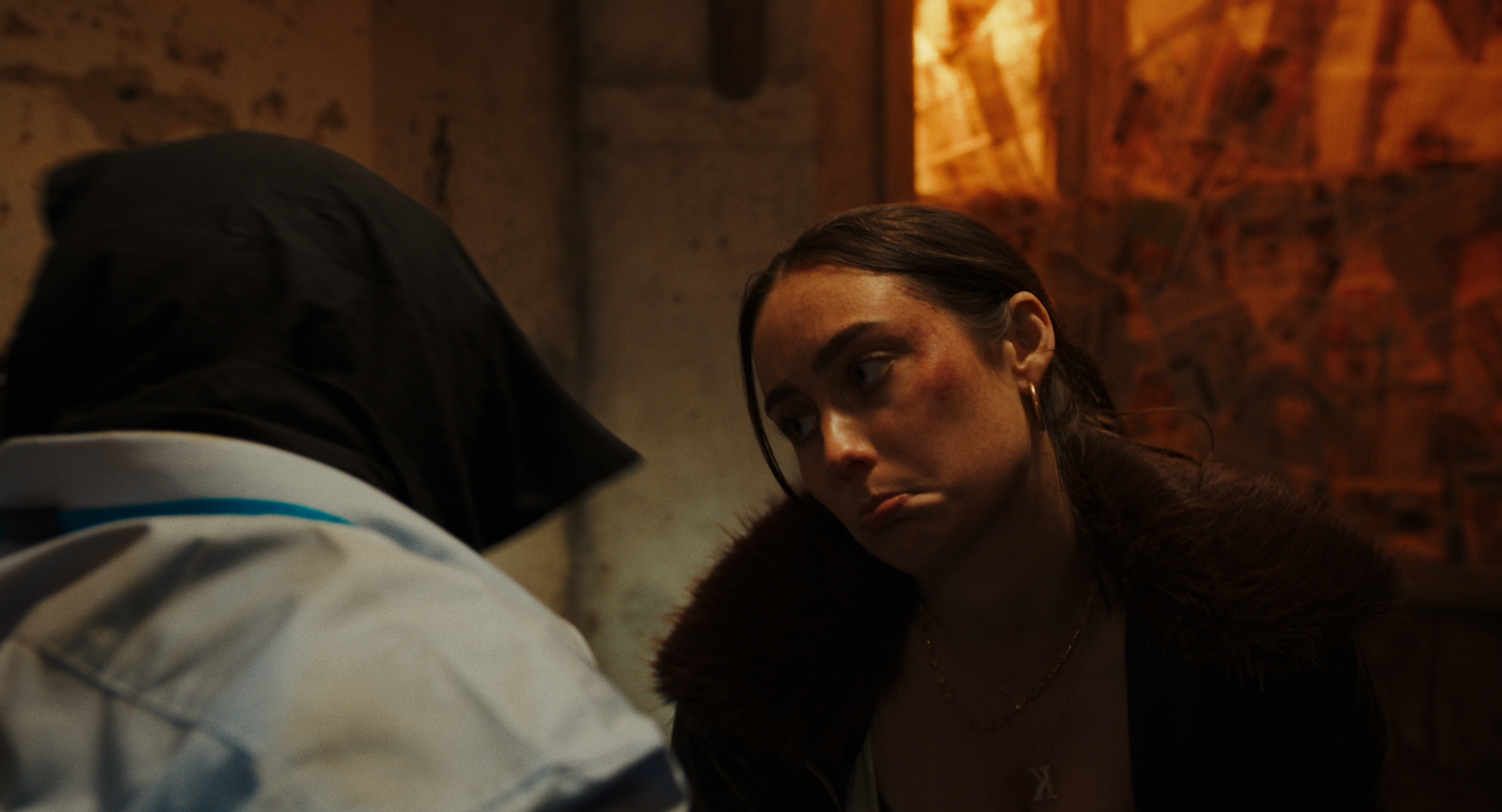 A woman with dark hair, wearing earrings and a fur collar, looks downward with a somber expression in a dimly lit room with brick walls.