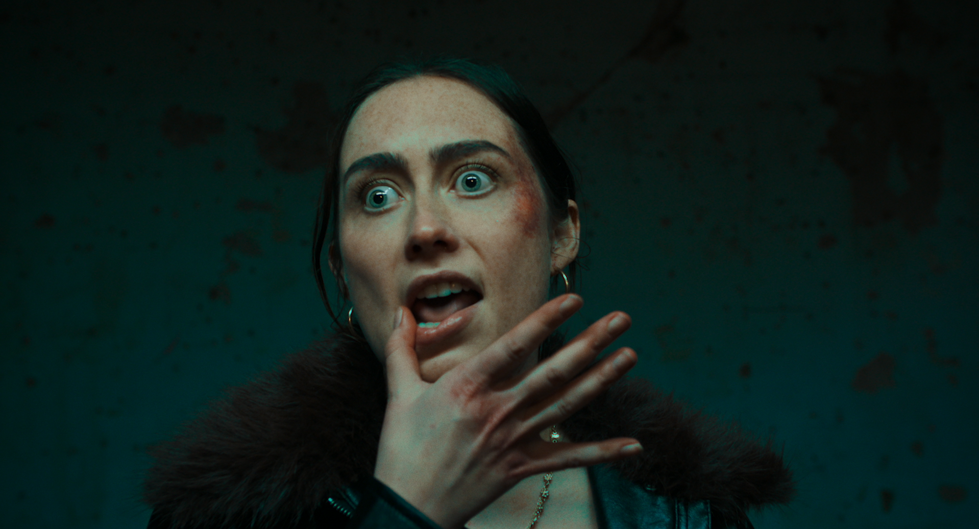 A woman with light skin and blue eyes, wearing a dark fur coat and hoop earrings, with feathered makeup on her face, holding her hand near her mouth with her fingers crossed. The background is dark and textured.