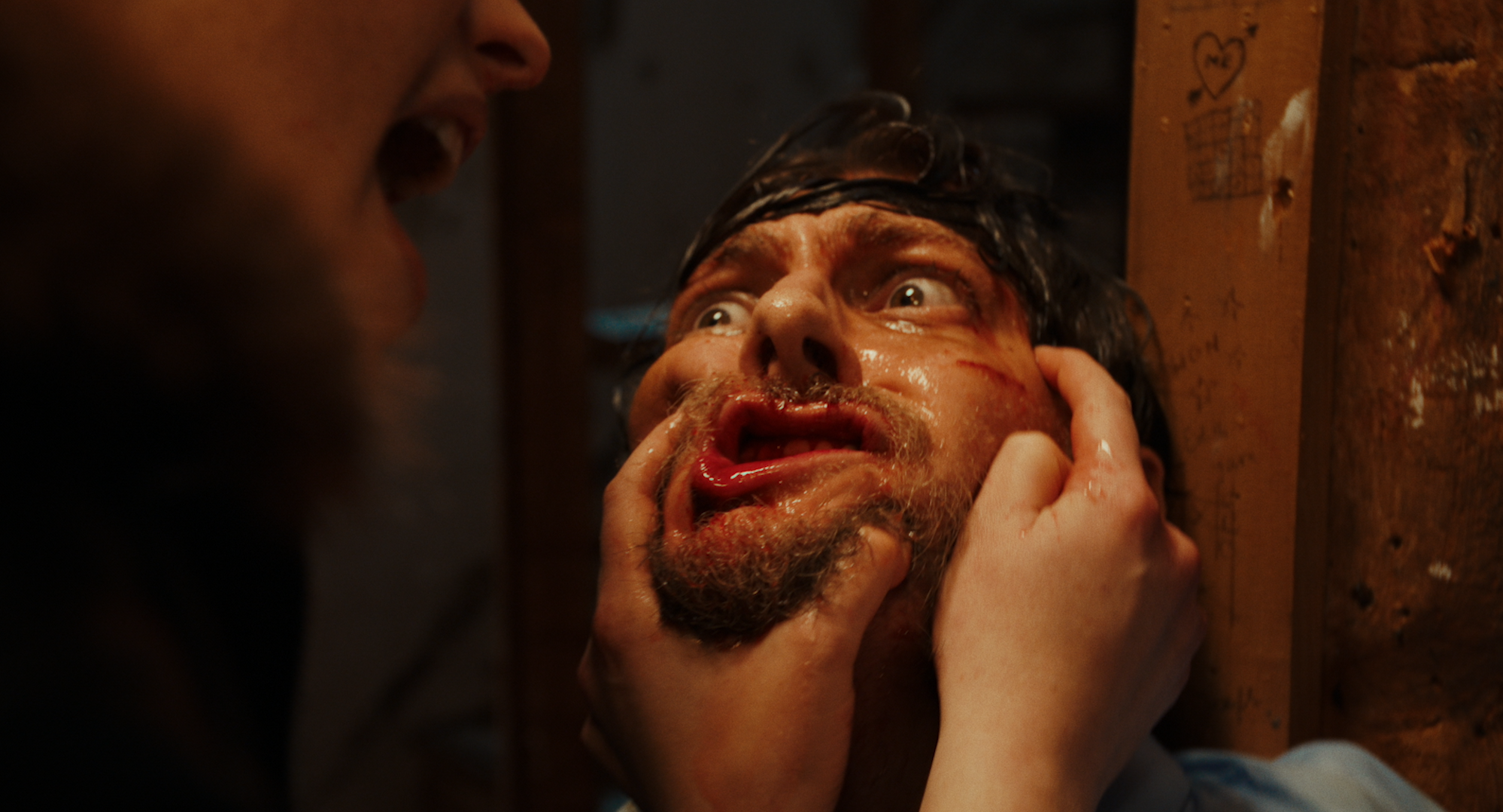 A person with a beard and wet hair appears distressed or surprised, while another person is holding their face and pressing it against a wooden surface with some doodles and writings on it.