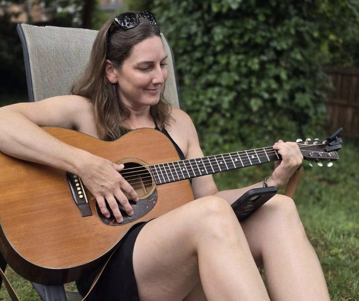 Person sitting outdoors playing an acoustic guitar, wearing sunglasses on their head, with greenery in the background.