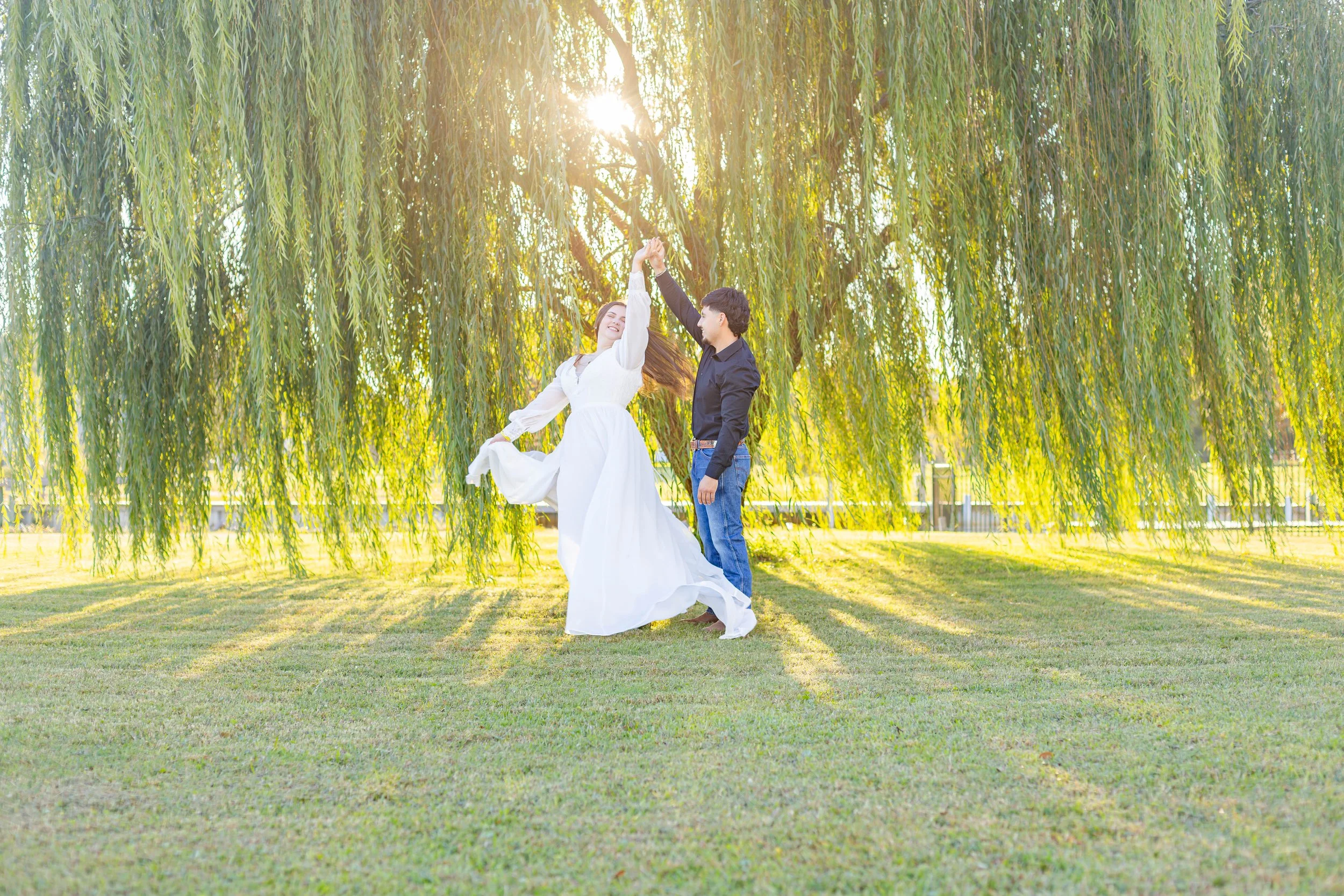 A couple dancing under a large willow tree in a park during sunset.